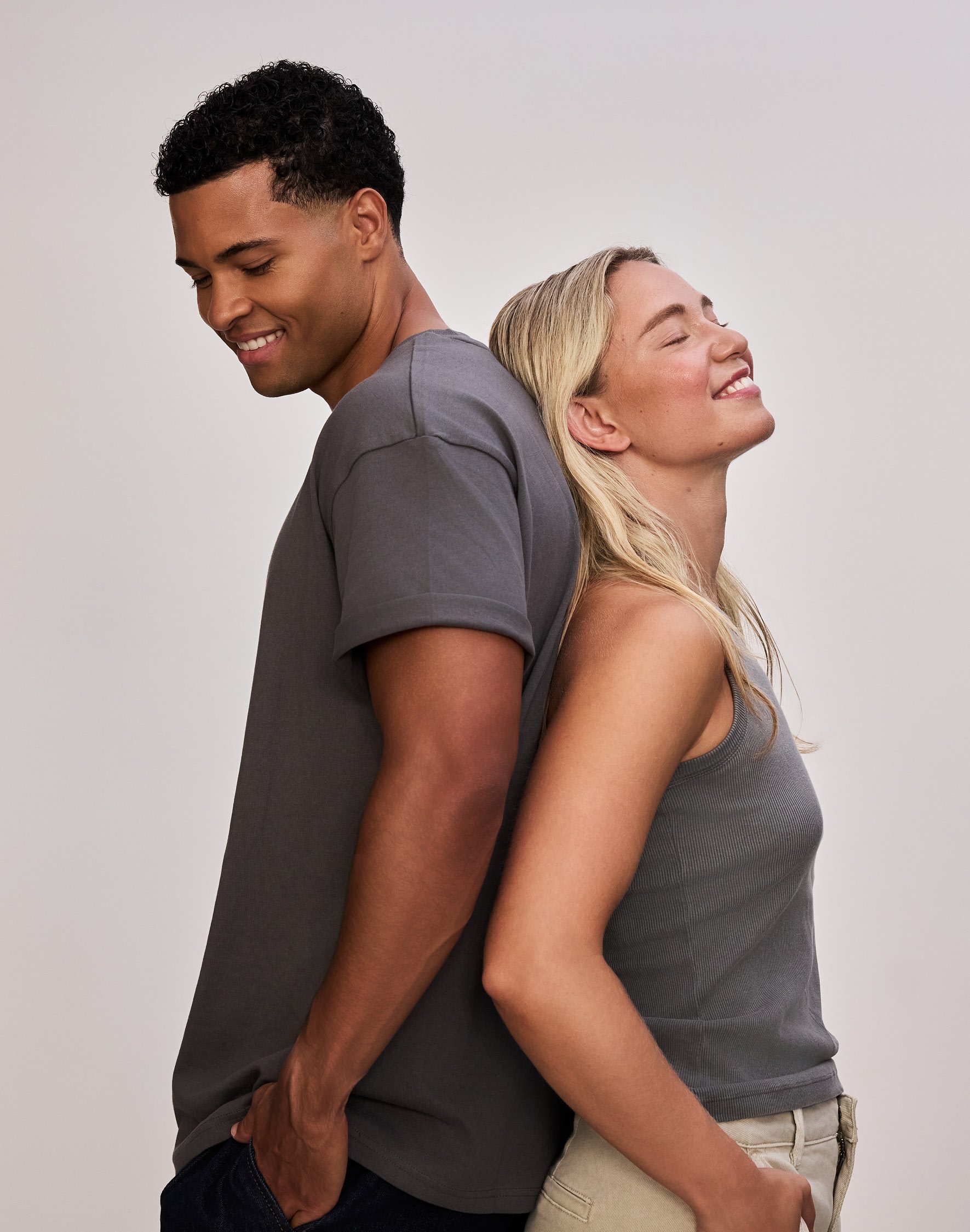 Ein Mann und eine Frau stehen Rücken an Rücken, lächeln und haben die Augen geschlossen. Er trägt ein graues T-Shirt, sie ein graues Top.