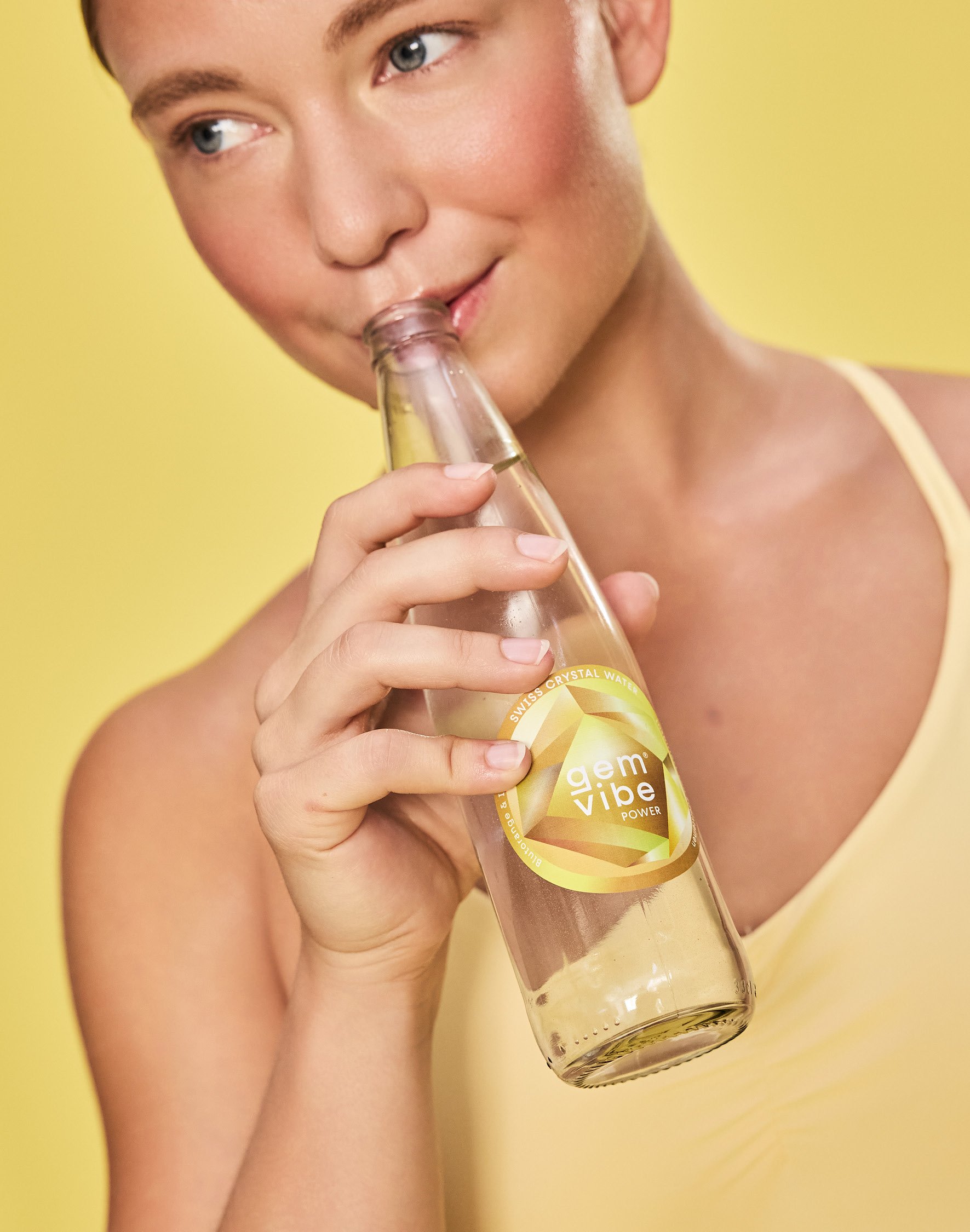 Nahaufnahme eines jungen Mannes, der ein Fläschchen Wasser mit dem Label "gem vibe" hält und an der Flasche nippt, vor einem gelben Hintergrund.