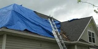 House with blue tarp covering the roof and a ladder leaning against it, indicating roofing or repair work in progress.