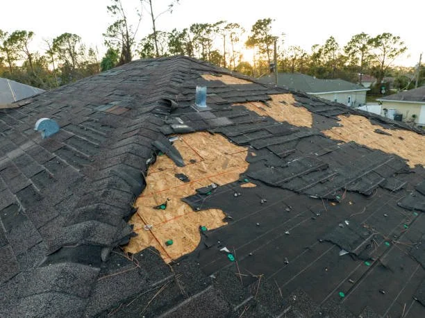 Roof with damaged and missing shingles, showing exposed plywood underneath, during sunset.