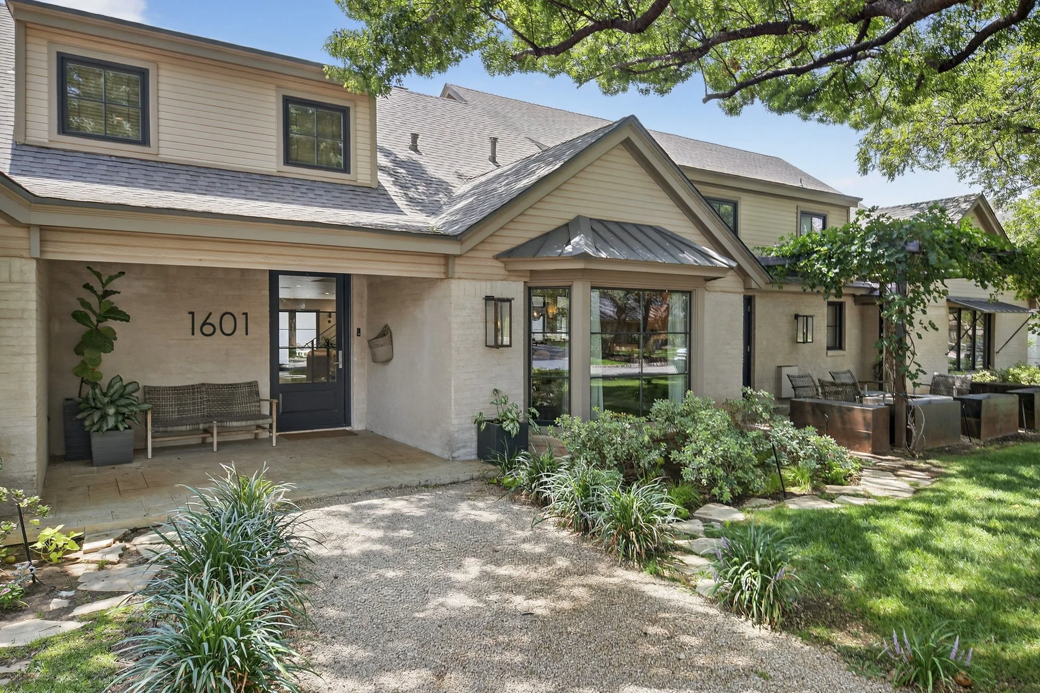 Front view of a modern house with beige siding, multiple windows, and a front porch with outdoor furniture, surrounded by a garden with various plants and a gravel pathway, with a large tree overhead and a clear blue sky.