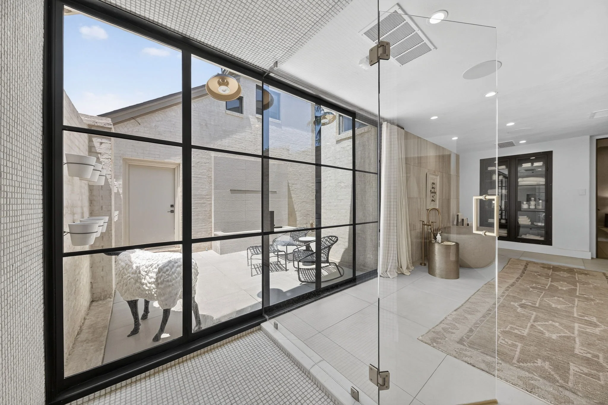 Modern interior with glass wall and outdoor patio view, featuring black-framed glass door, white textured wall, and light-colored furniture.