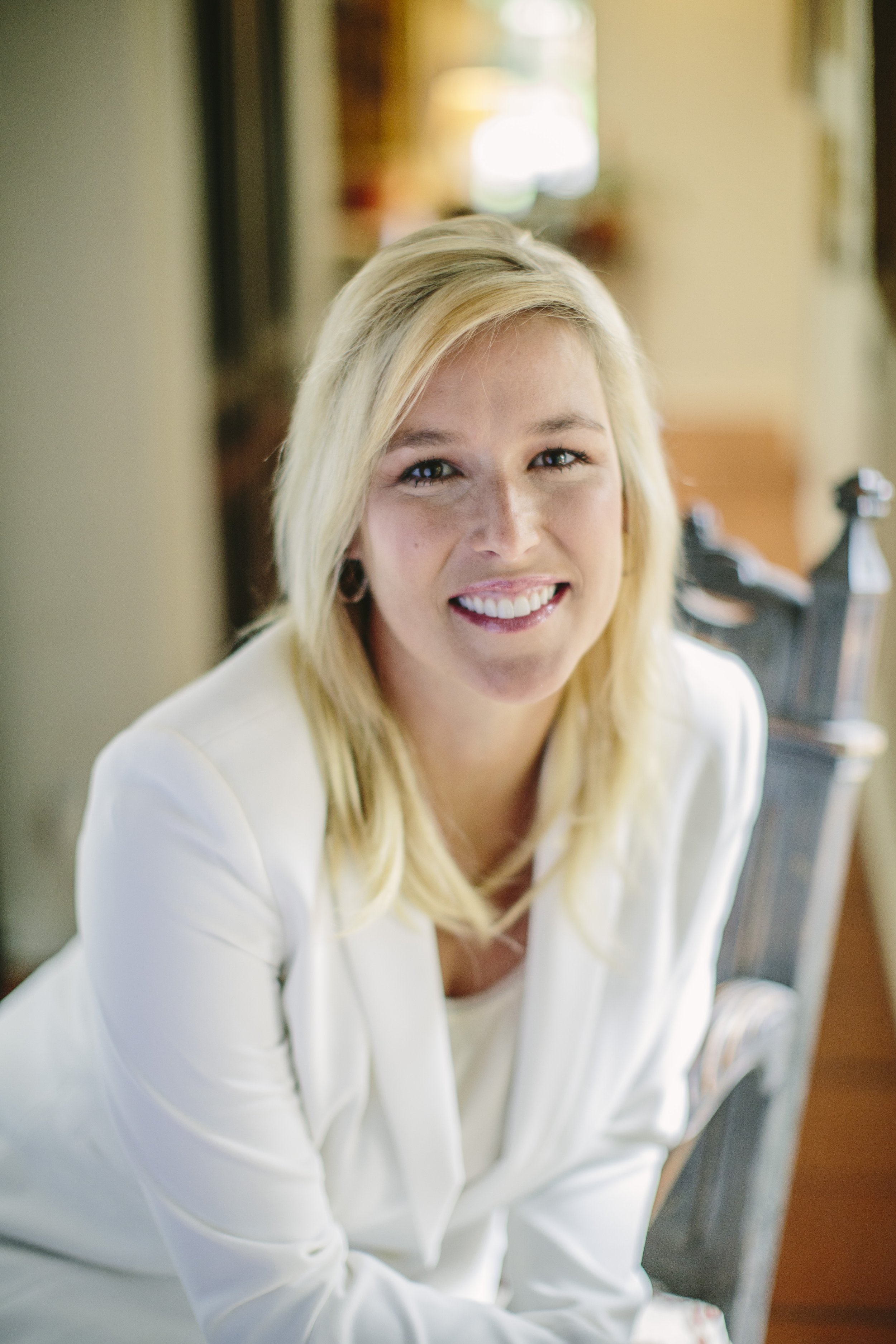 A smiling woman with blonde hair sitting indoors near a window, wearing a white blazer.