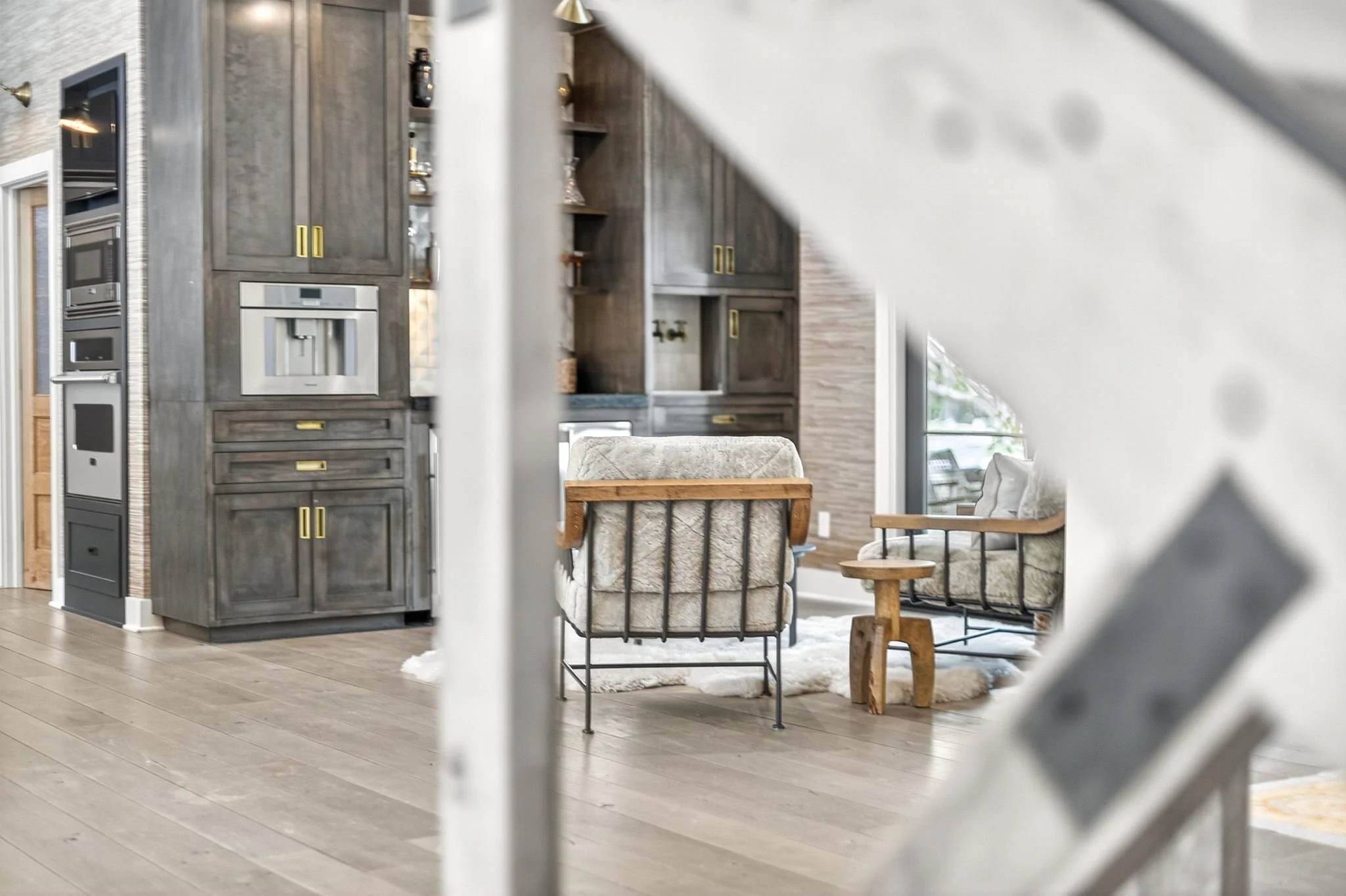 Interior view of a modern home showing a kitchen with gray cabinetry, built-in stainless steel appliances, and a living area with chairs, a small table, and large windows.