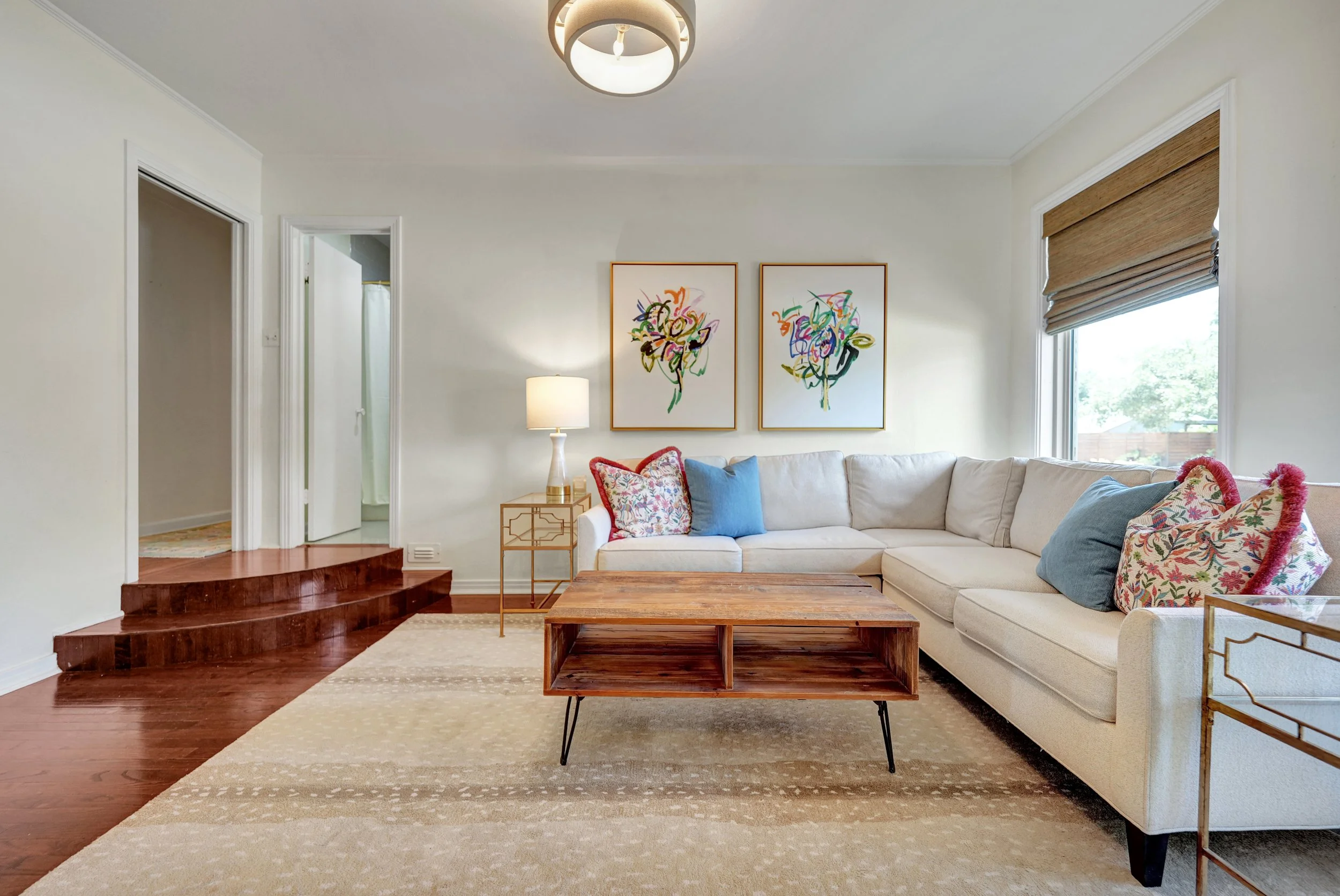 Living room with white sectional sofa decorated with colorful pillows, a wooden coffee table, a side table with a lamp, two framed abstract art pieces, a window with bamboo blinds, a ceiling light, and a light-colored area rug over dark wood flooring.