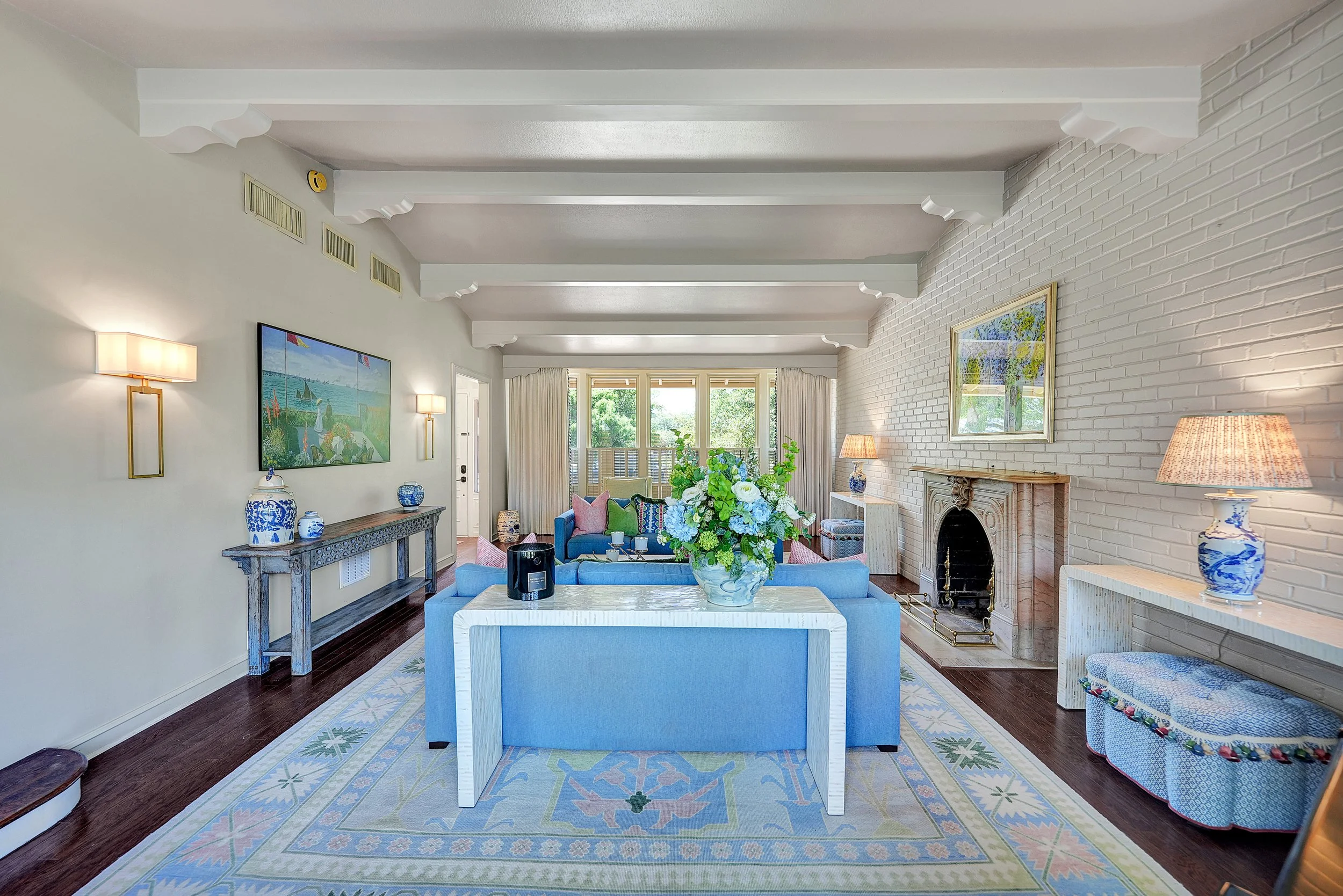 Living room with white brick fireplace, blue sofas, floral arrangement on white table, artwork on walls, table lamps, and large windows with curtains.
