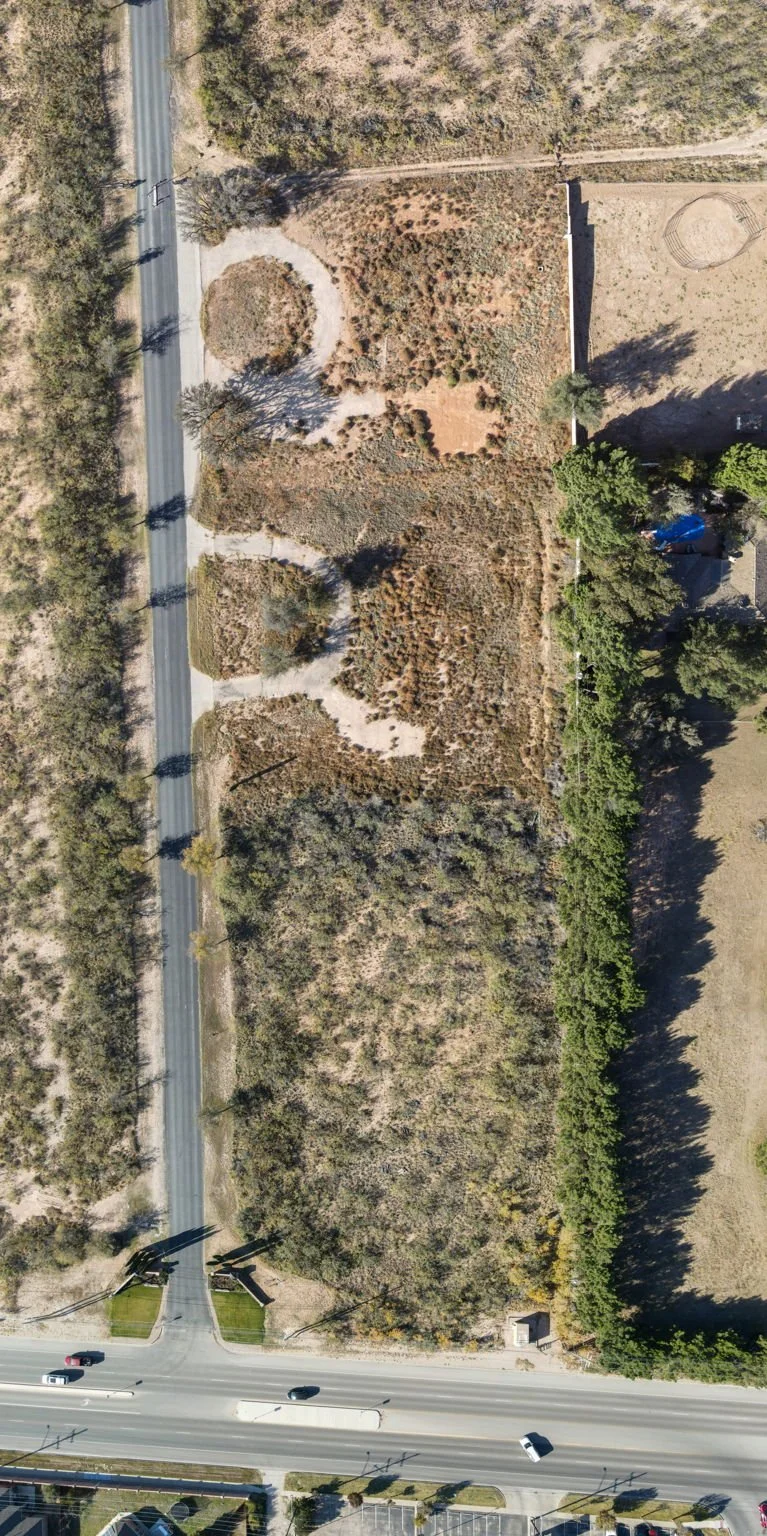 Aerial view of a lot with sparse vegetation and trees, bordered by roads on the left and bottom, with a small structure near the bottom right corner.