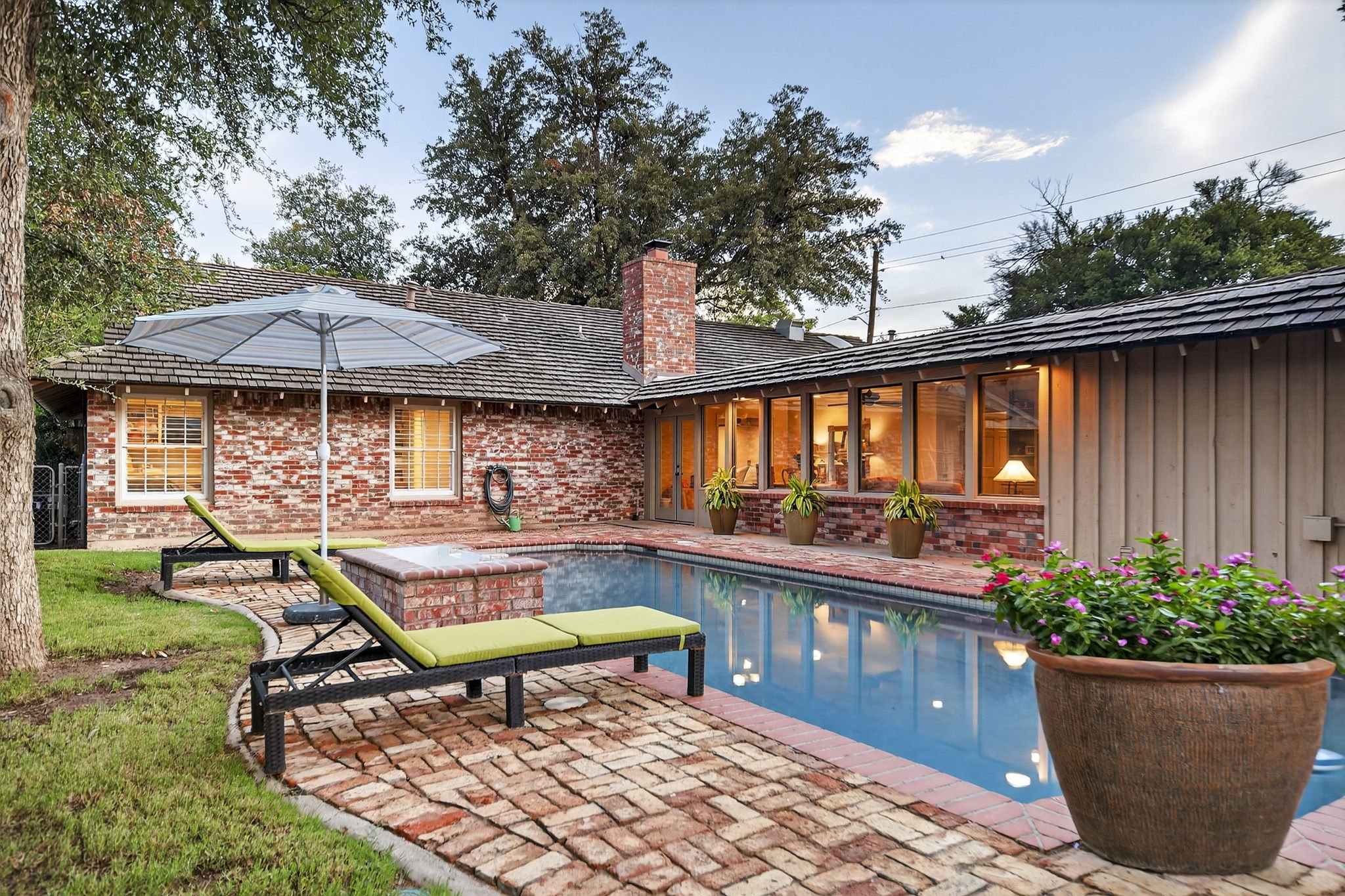 Backyard view with a swimming pool, lounge chairs, a large potted plant, brick patio, house with brick and siding exterior, large windows with interior lights visible, umbrella, trees, and a blue sky with clouds.