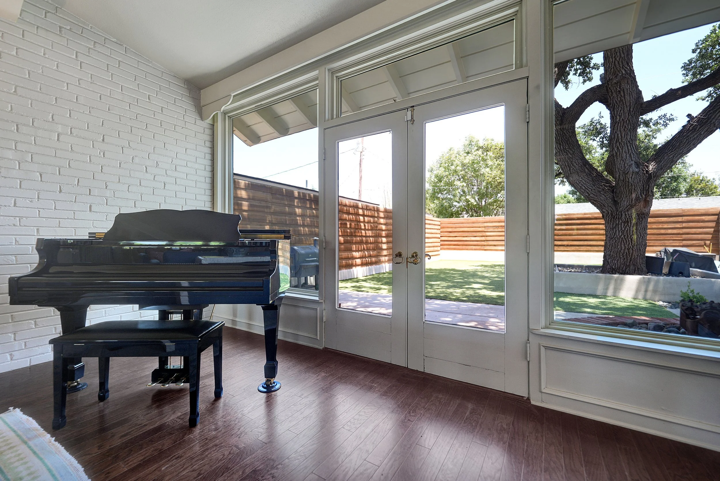 Living room with a large windowed door leading to a backyard with a tree and a wooden fence, a black grand piano, and hardwood floors.