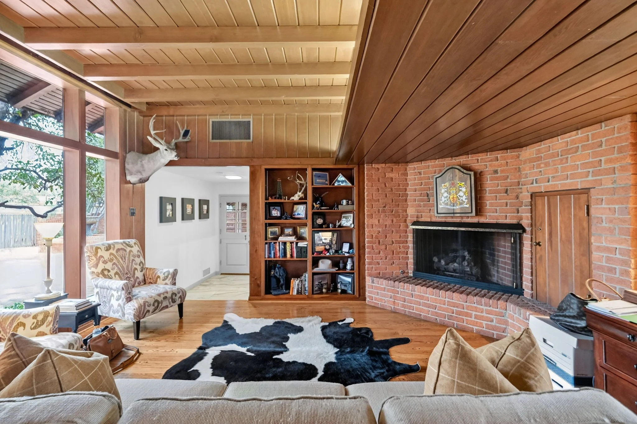 Living room with a brick fireplace, wooden ceiling, large window, and various furniture and decorations.