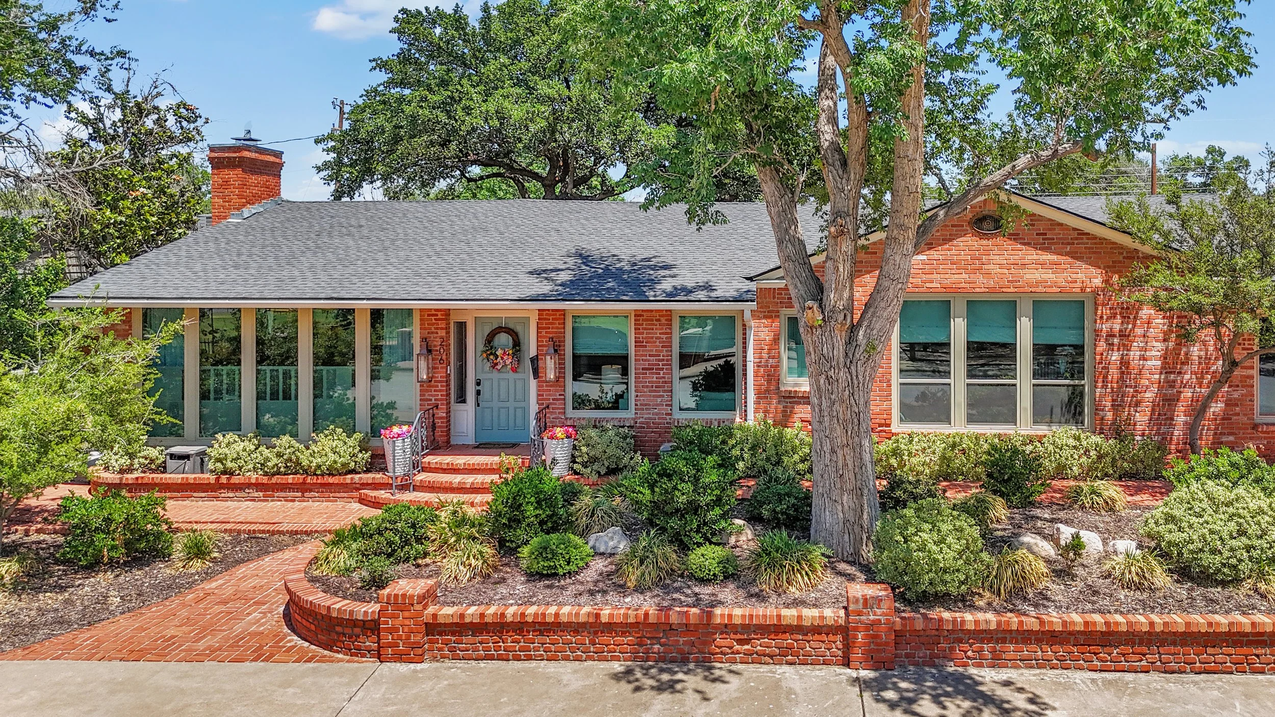 Front view of a red brick house with a porch, surrounded by lush landscaping and trees, under a blue sky.