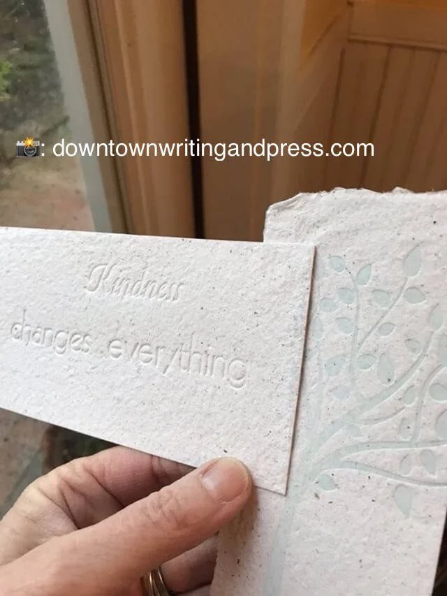 Close-up of a hand holding a textured, off-white card with the embossed message 'Kindness changes everything' and an embossed decorative plant design on the right side, indoors with wooden paneling in the background.