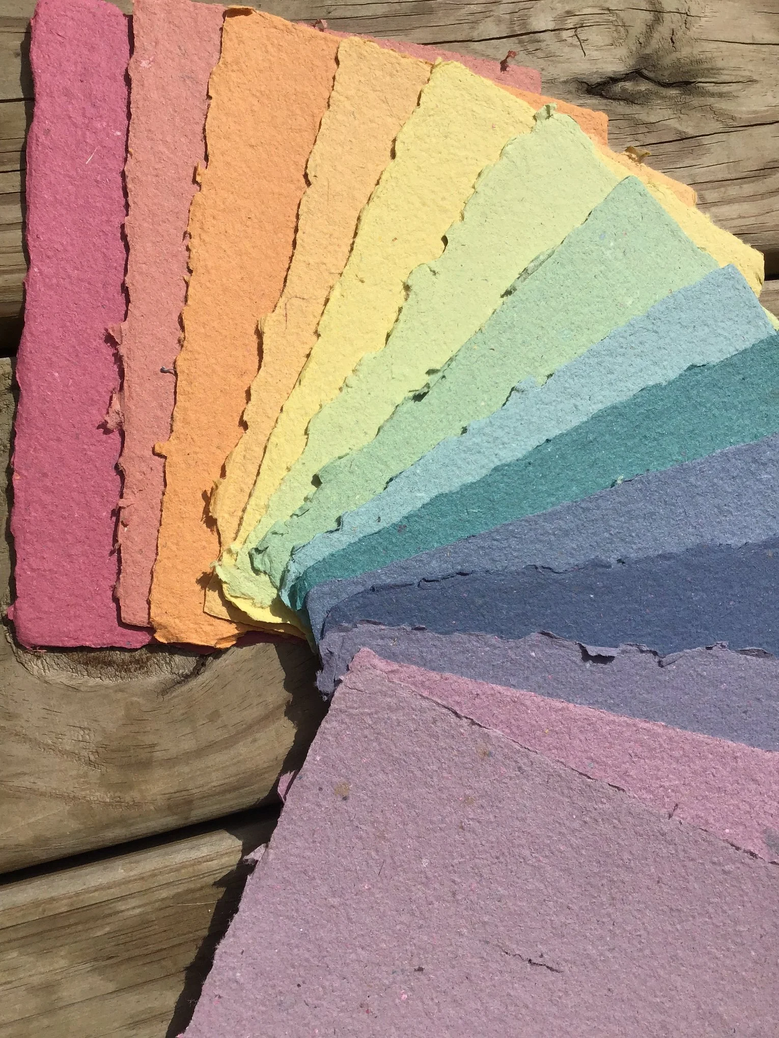 A gradient of deckled edge handmade paper sheets fanned out in order: red, orange, yellow, spring green, light and dark teal, light and dark blue, and light and dark purples.