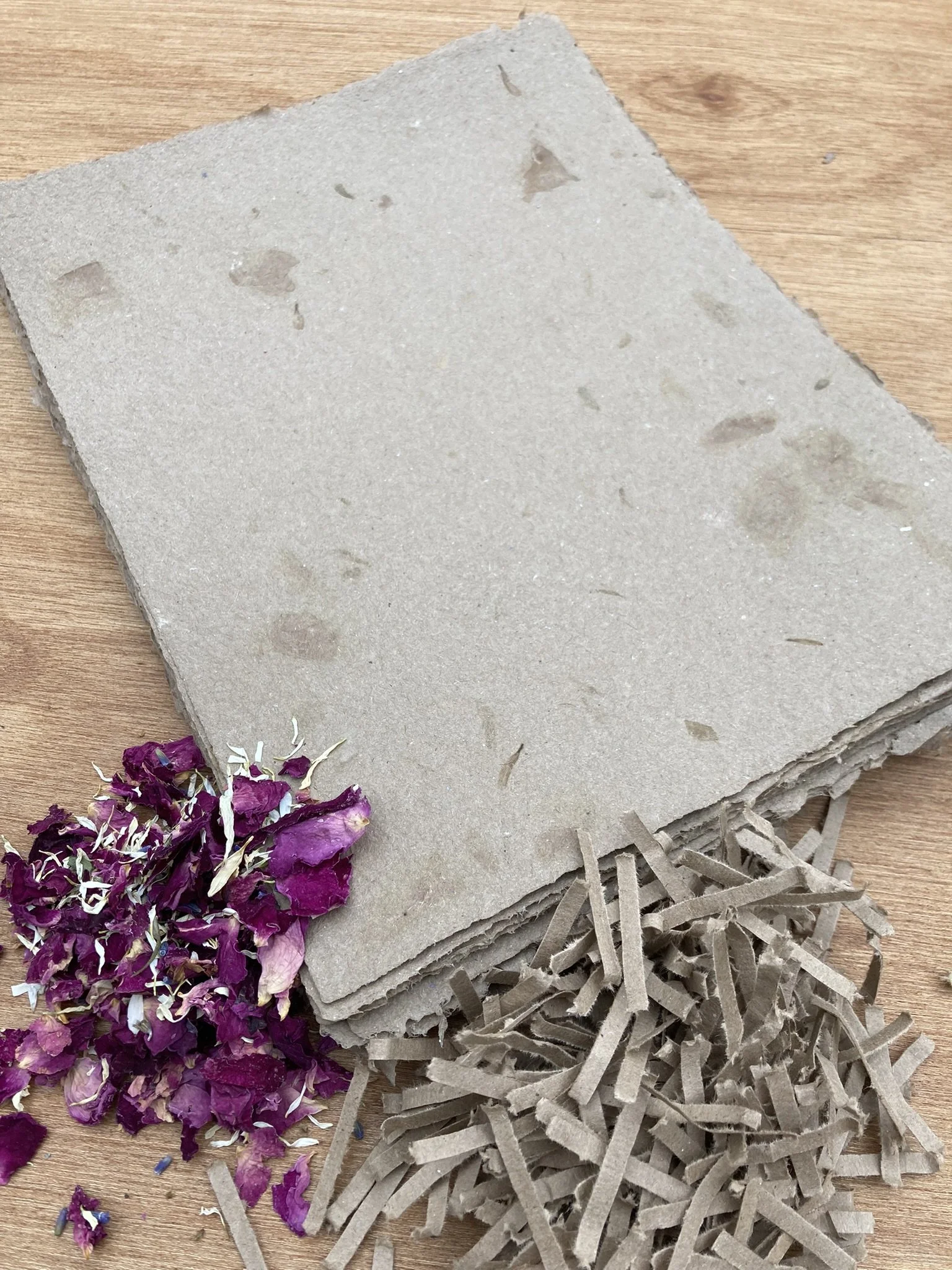 a stack of brown paper with deckled edges and rose petal flower inclusions.  An attractive pile of rose petals and shredded cardboard are draped over the stack, showing the paper's origin fibers.