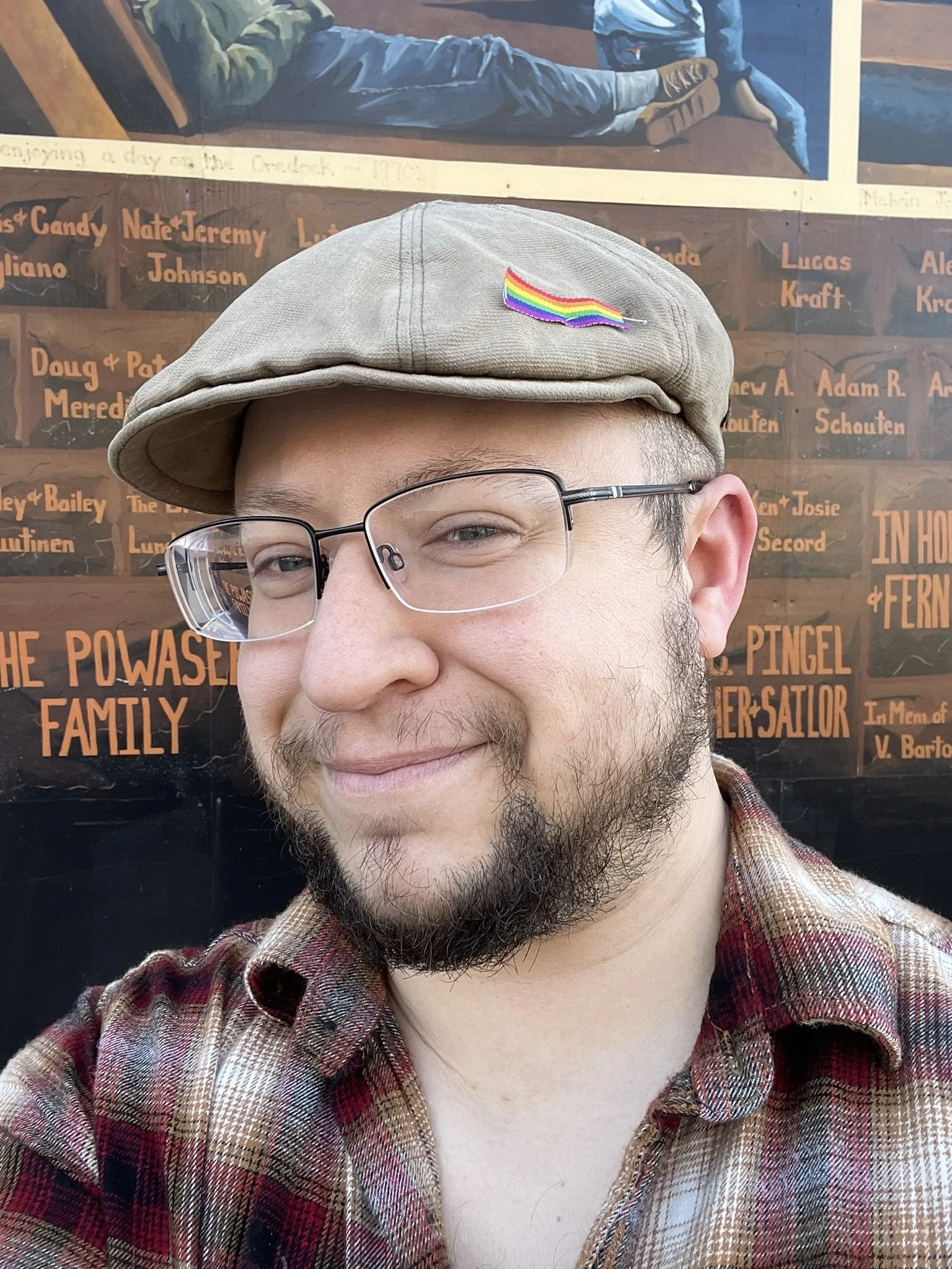 An image of a nonbinary person in a burgundy and tan plaid button up shirt and a flat cap with a small rainbow ribbon pilled to one side.  They are wearing black half rim glasses and have a full beard of brown hair.