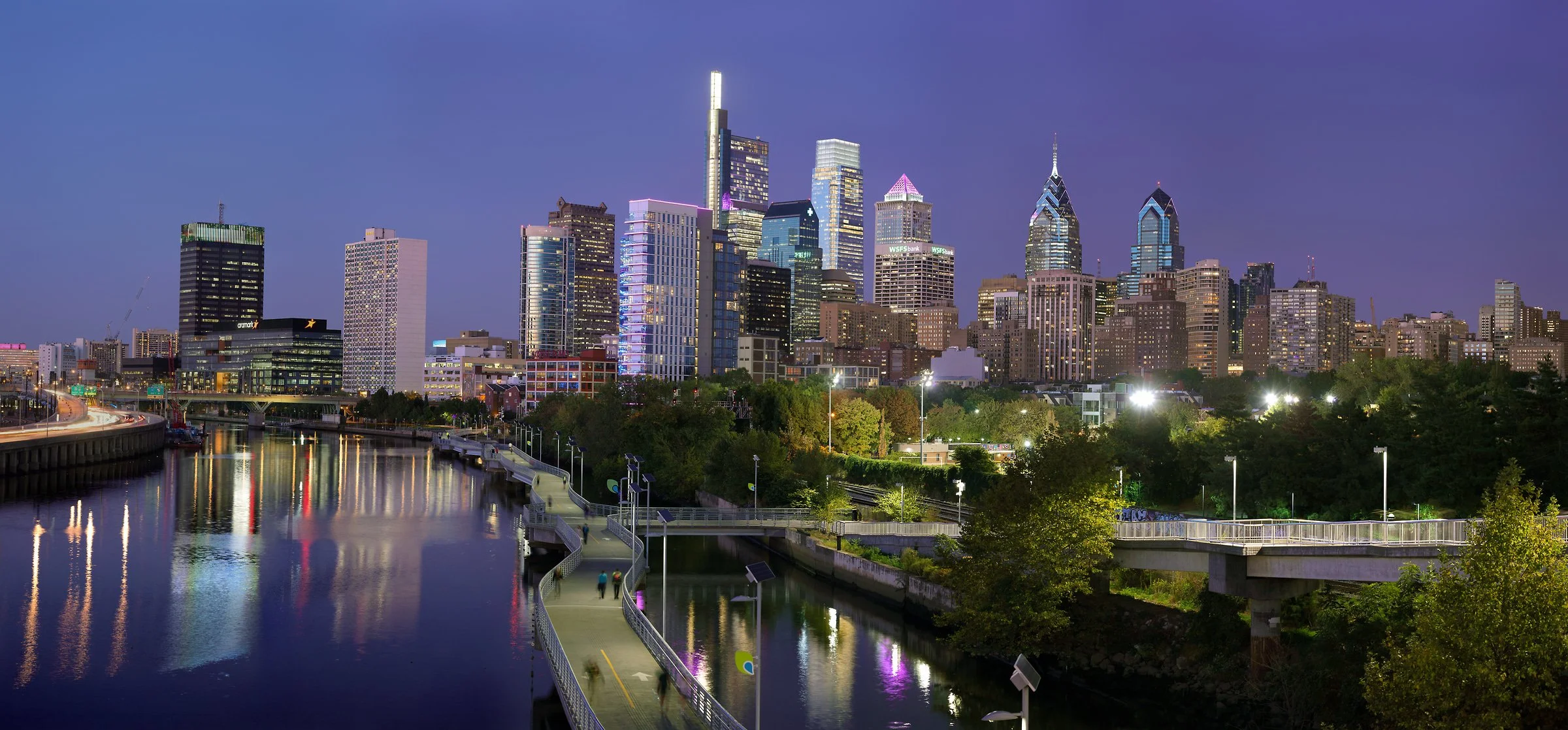 Night skyline of a city with tall skyscrapers illuminated, reflecting on the river below, with a walking and biking trail along the water and trees in the foreground.