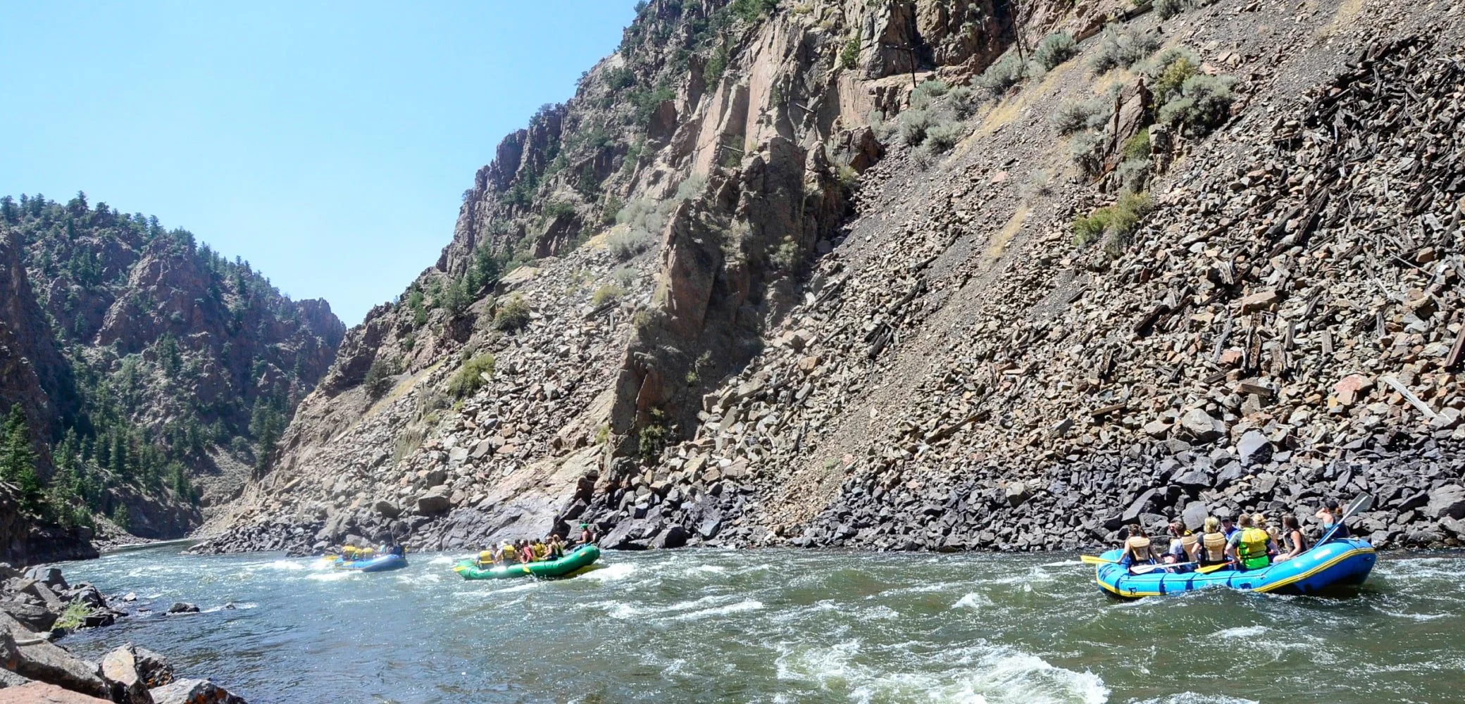 People rafting down a river surrounded by steep rocky cliffs and forested mountains under a clear blue sky.