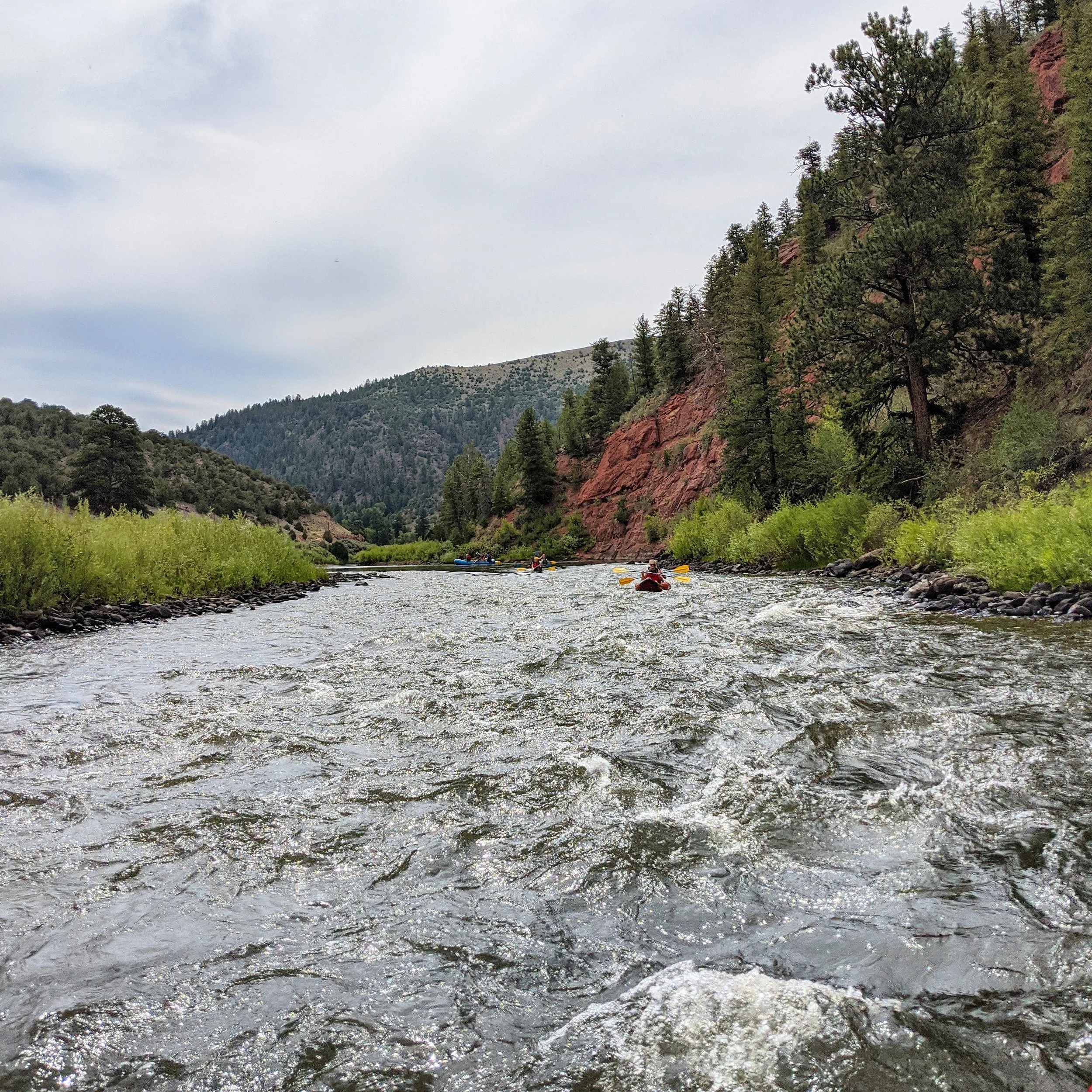 A river flowing through a mountainous landscape with green trees and rocky red cliffs under a cloudy sky.