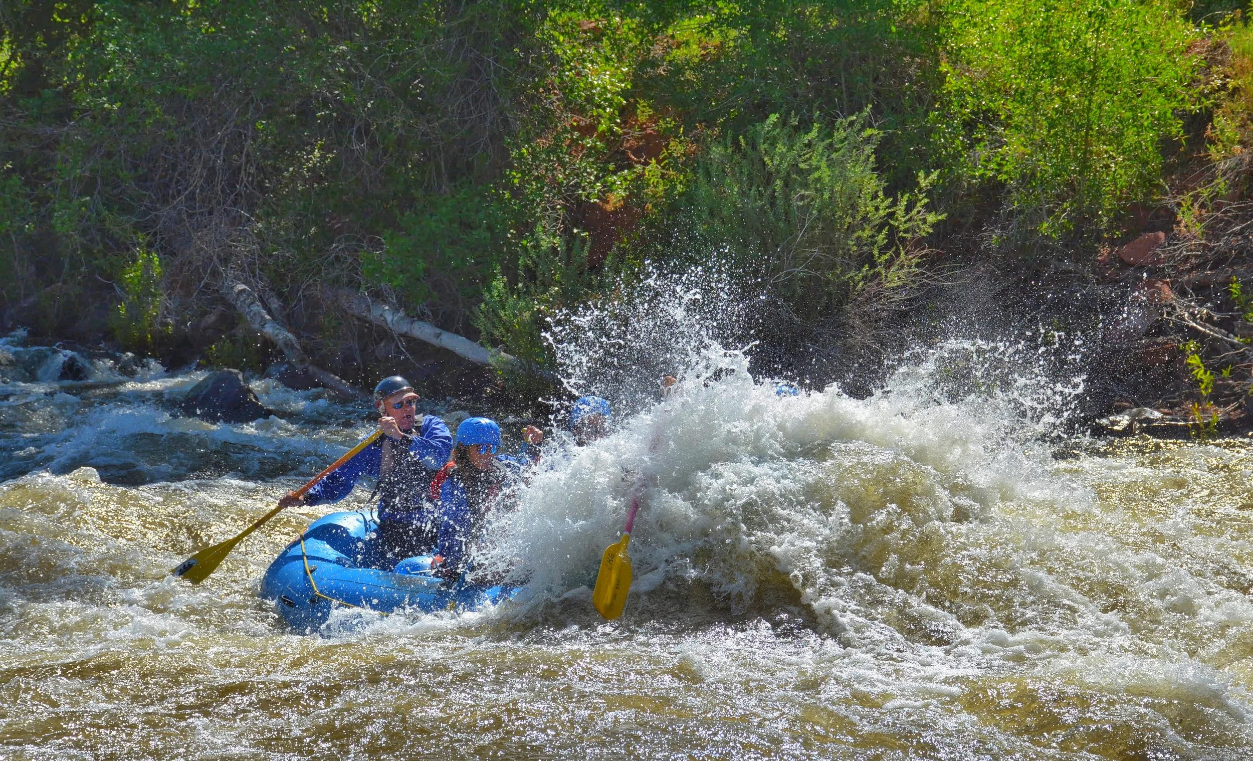 People rafting on a river surrounded by greenery, with water splashing.