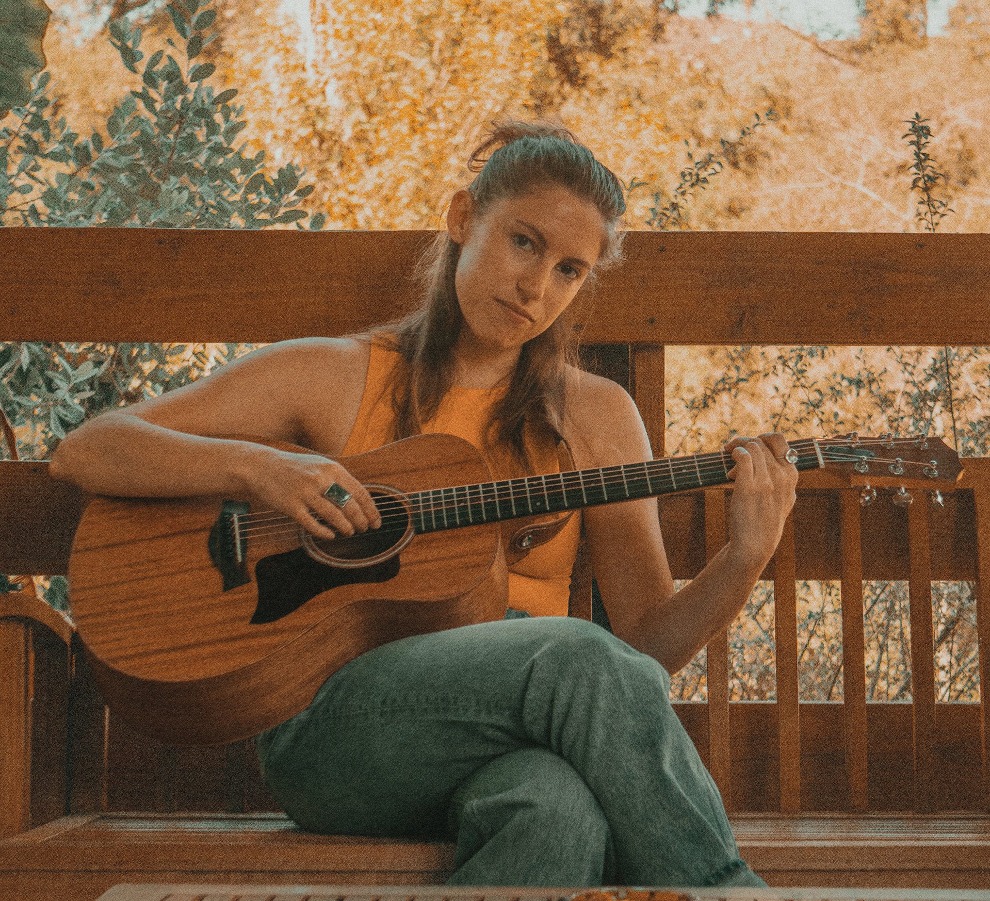 A person with long brown hair, wearing a yellow top, playing an acoustic guitar, seated outdoors with a background of trees and leaf-covered ground.