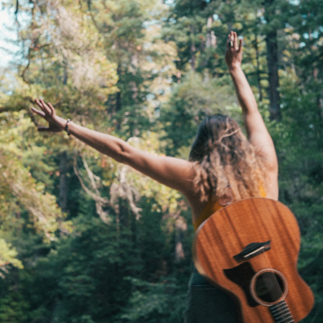 A woman with curly hair playing an acoustic guitar outdoors in a forest with green trees, wearing a sleeveless top, with her arms raised in the air.