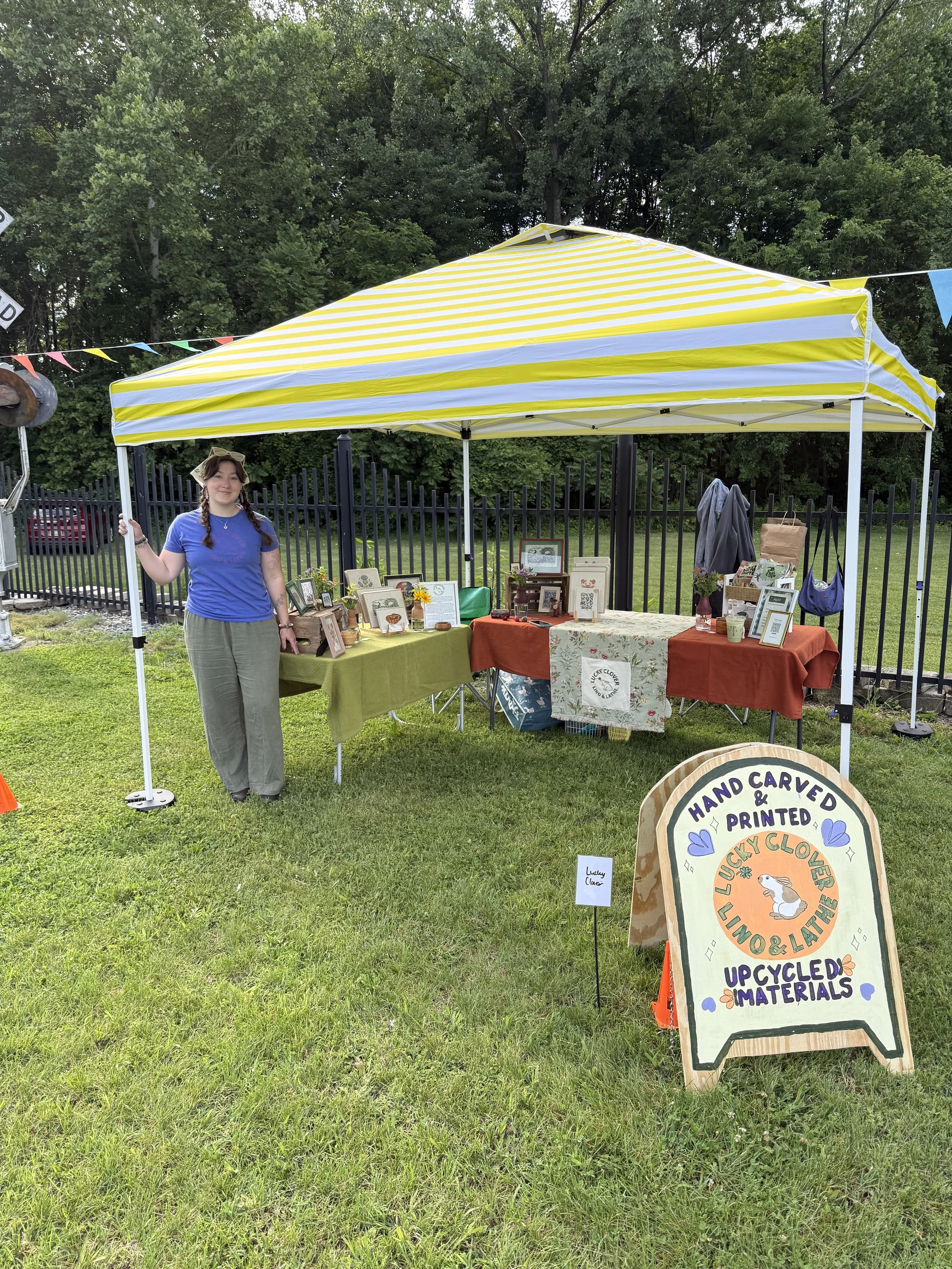 A woman standing under a yellow and white striped canopy at an outdoor craft booth, with tables displaying handmade items, signs, and a fenced grassy area in the background.