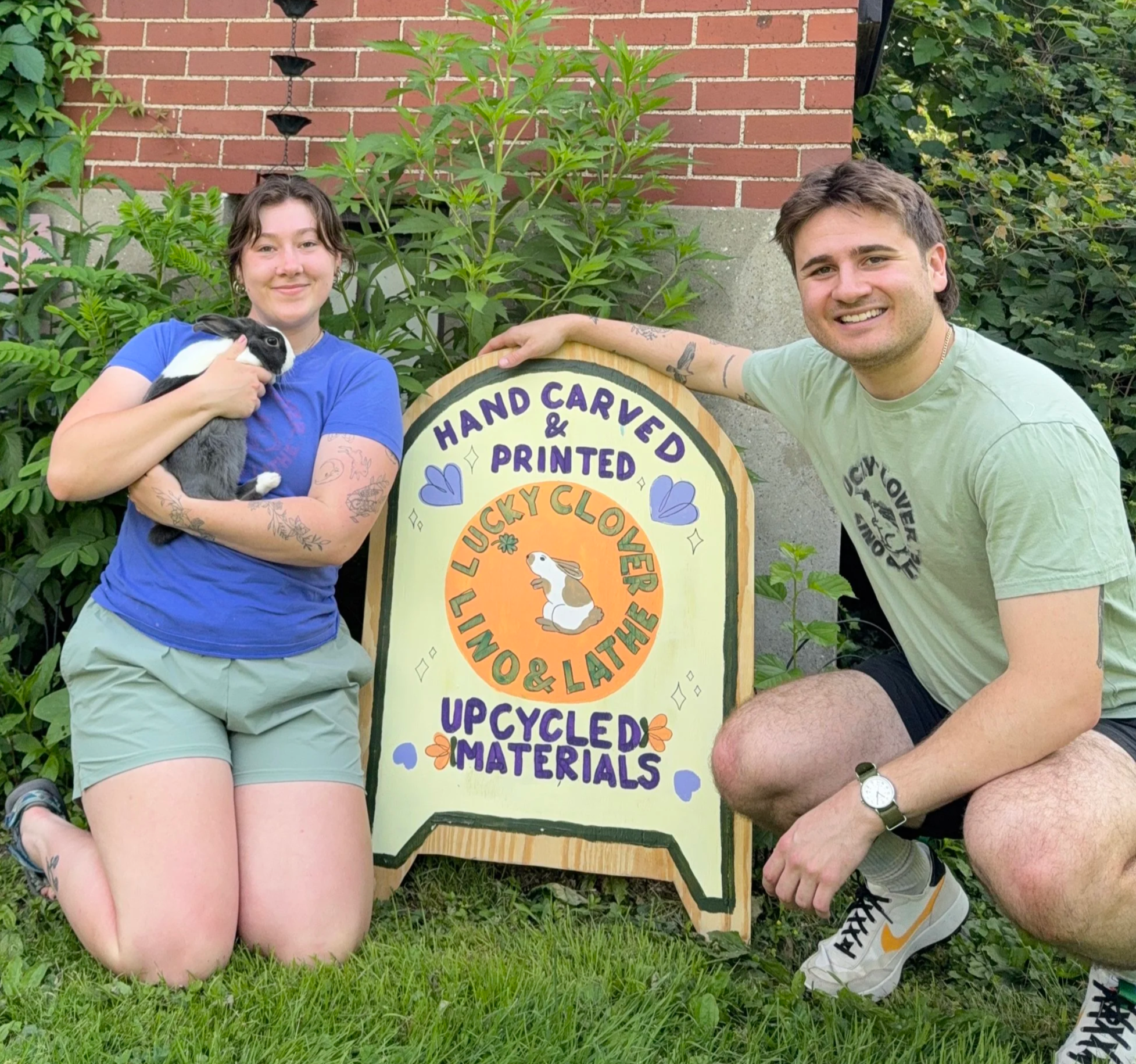 Two people kneeling on grass in front of a sign that reads "Hand Carved & Printed Lucky Clover Upcycled Materials." One person is holding a rabbit, and the other has tattoos on her arm. There are plants and a brick wall in the background.