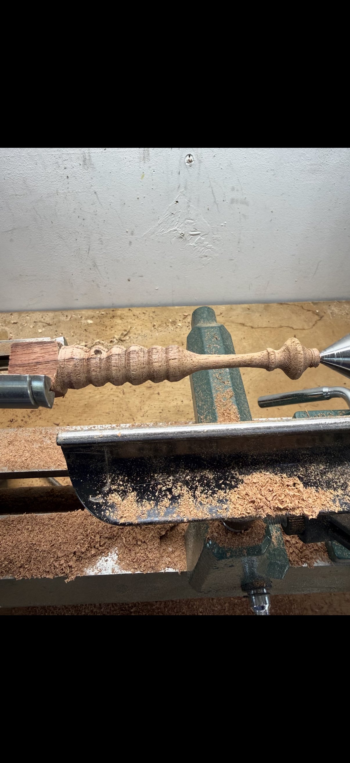 A woodworking lathe machine shaping a wooden spindle with wood shavings collected around it.