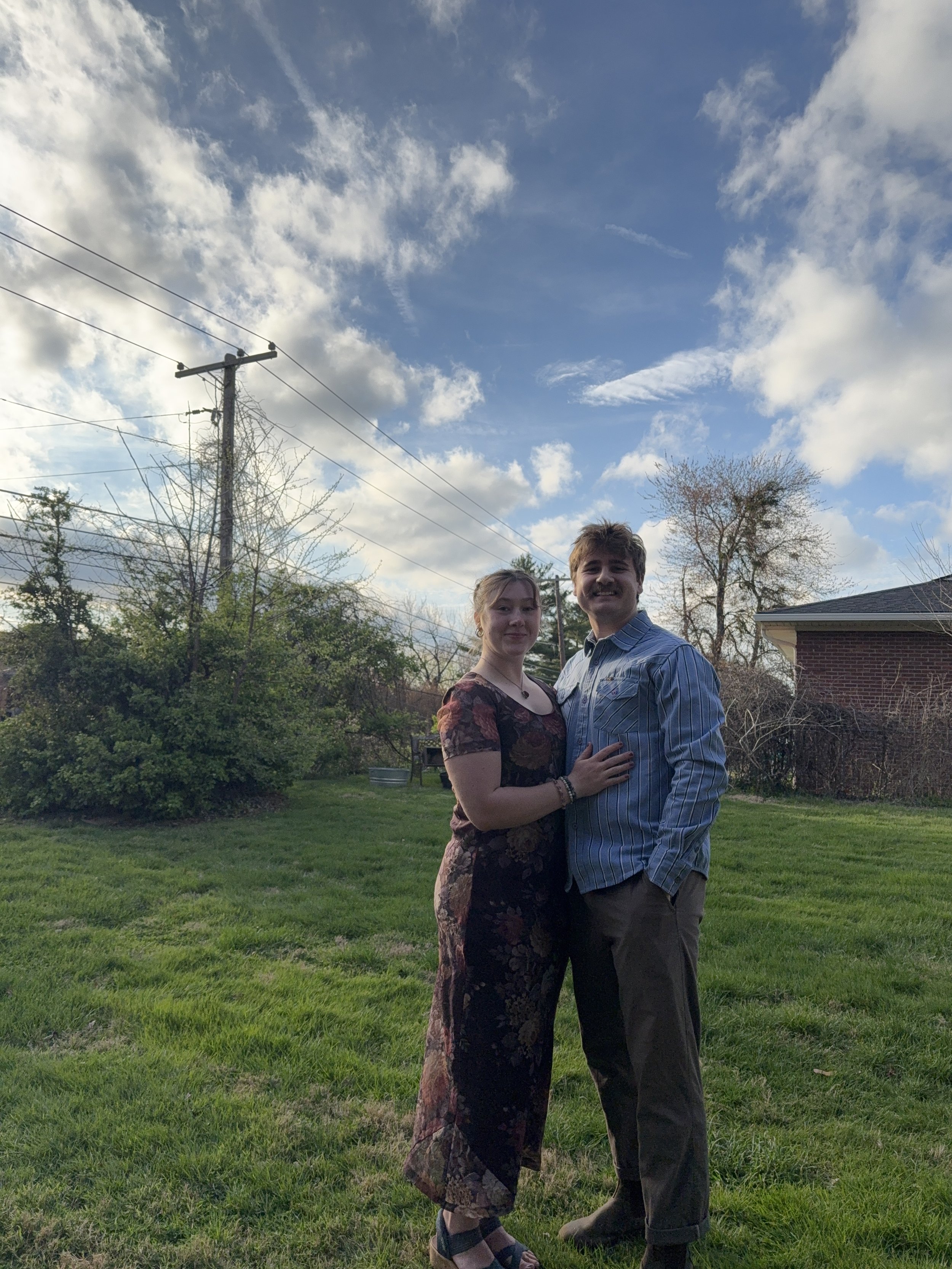 A man and a woman standing outdoors on a grassy lawn, with trees, a brick house, and a utility pole with power lines in the background. The man is smiling and wearing a striped shirt, while the woman has a neutral expression and is wearing a floral dress.