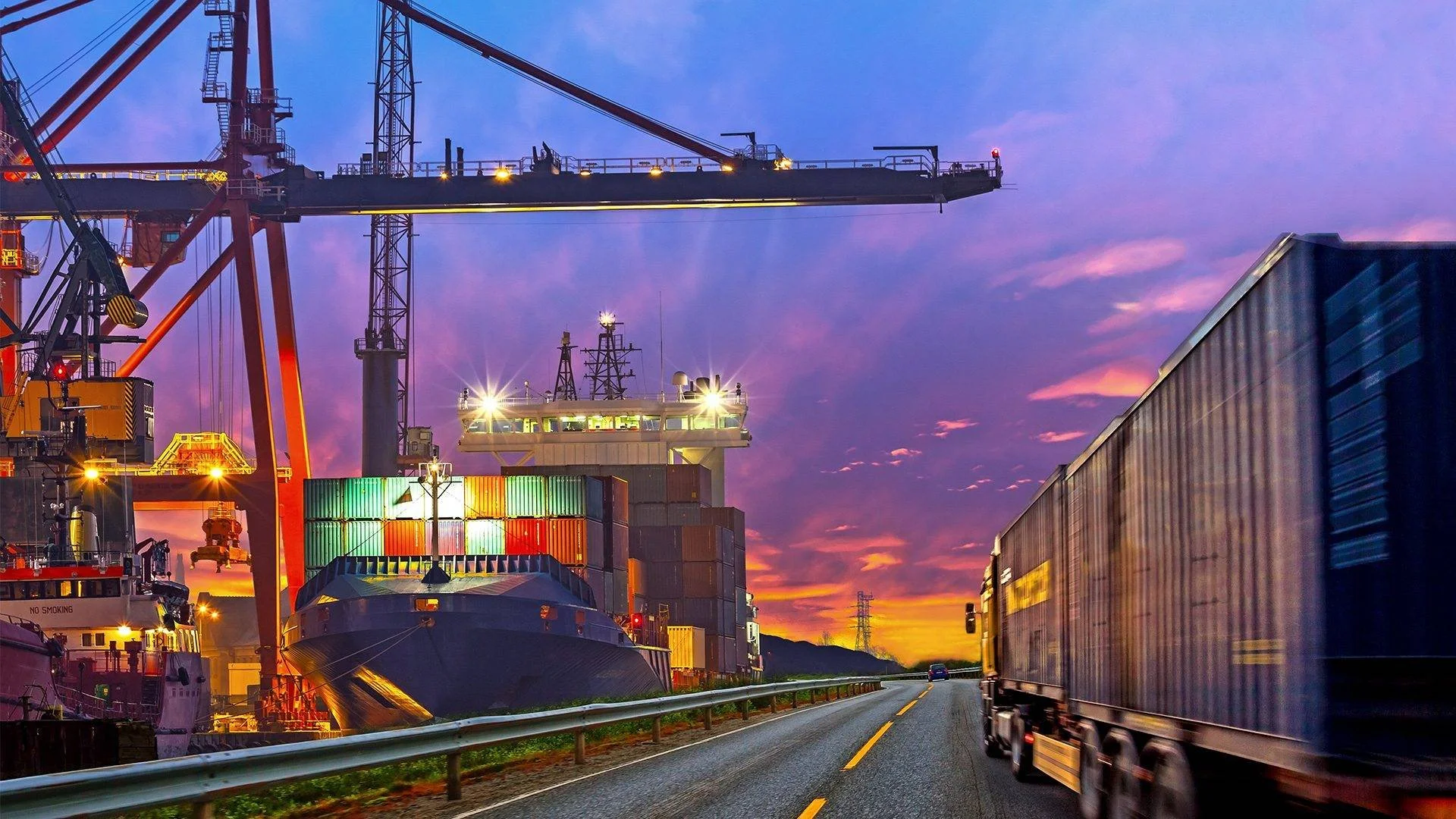 A busy shipping port at sunset with containers, cargo ships, cranes, and trucks on a highway.
