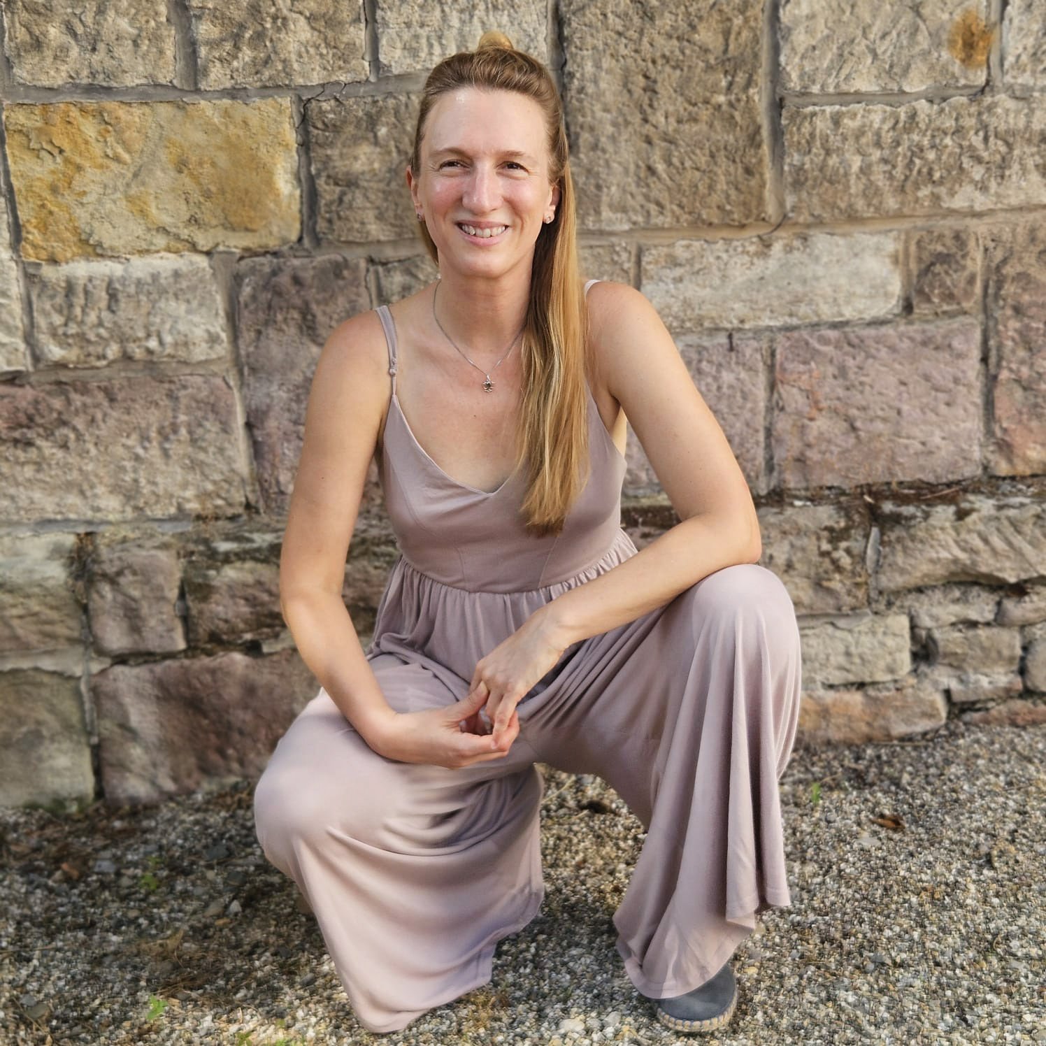 Eine sympathisch lachende Frau mit langen blonden Haaren in einem beigefarbenem sommerlichen Overall sitzt vor einer Steinmauer.