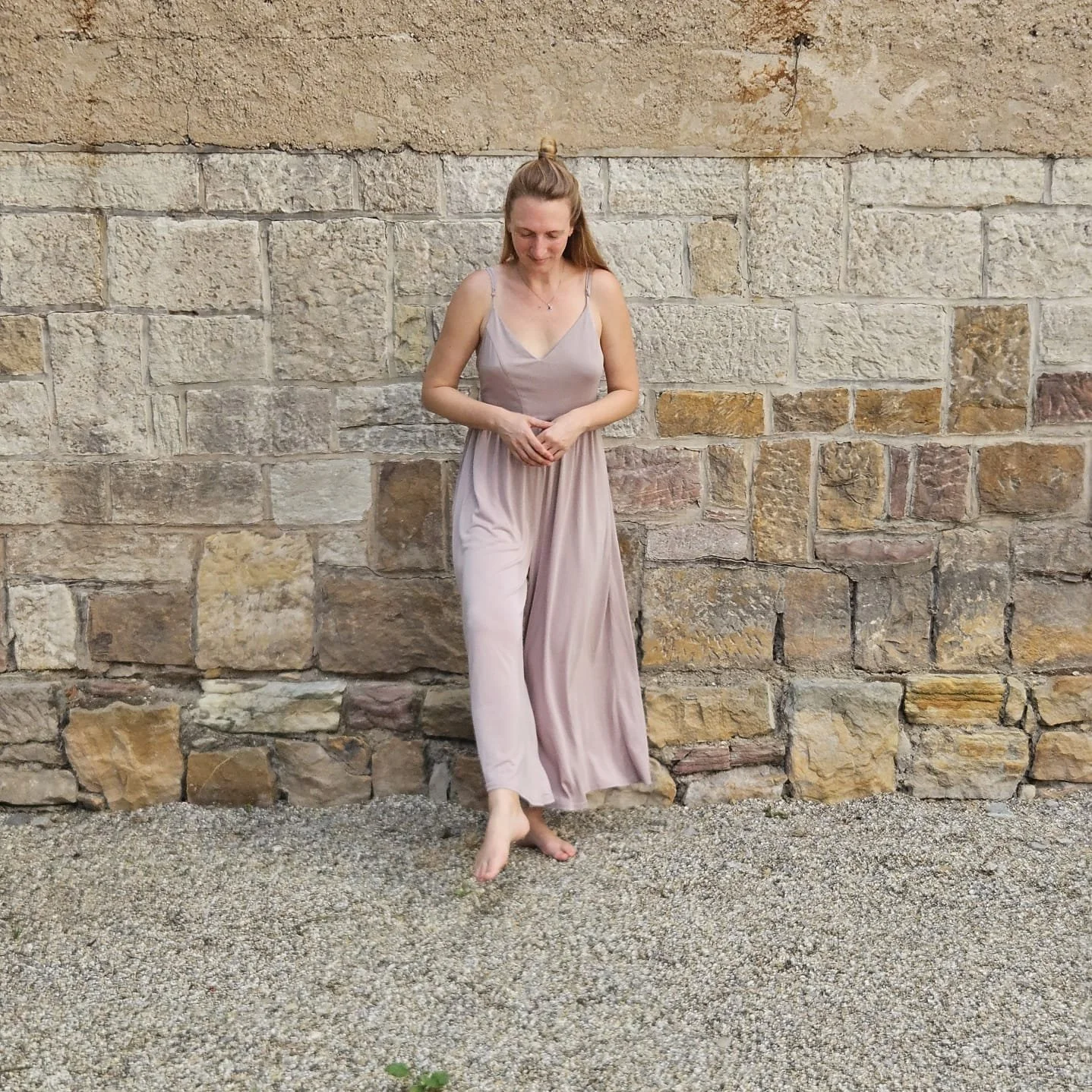 Eine junge Frau in einem fließenden, hellrosa Kleid, barfuß, steht vor einer Steinmauer, lachend mit nach unten gerichtetem Blick, auf Kieselsteinen stehend.
