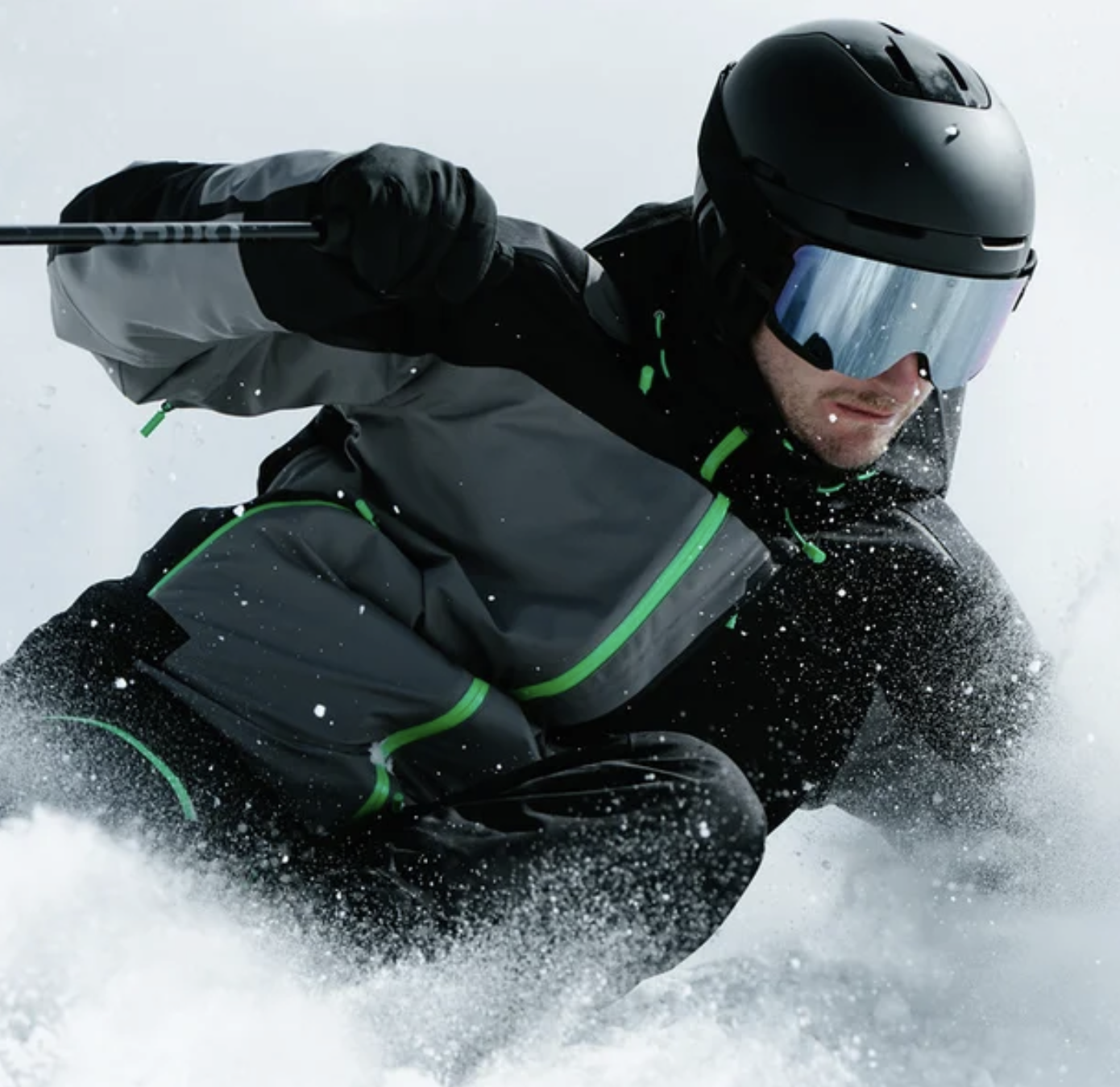 Ein Skifahrer trägt eine schwarze Helm mit Visier, schwarze und graue Bekleidung mit grünen Akzenten, und fährt auf Schnee, wobei er sich nach einem Sturz befindet.