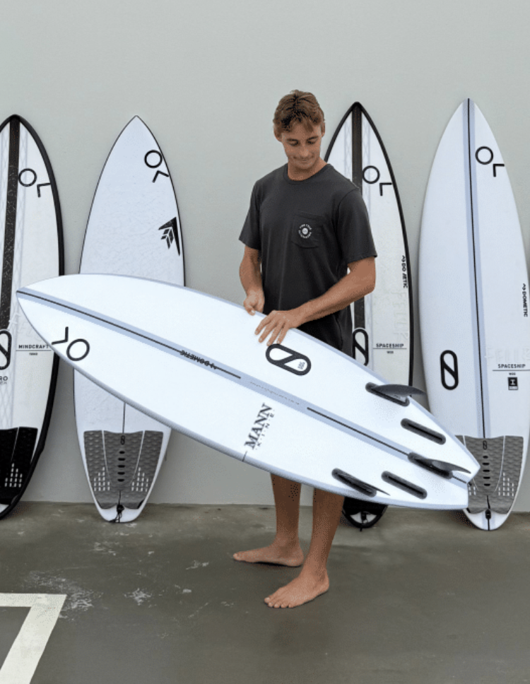Junger Mann hält ein weißes Surfbrett in einem Raum mit mehreren weiteren Surfboards im Hintergrund.