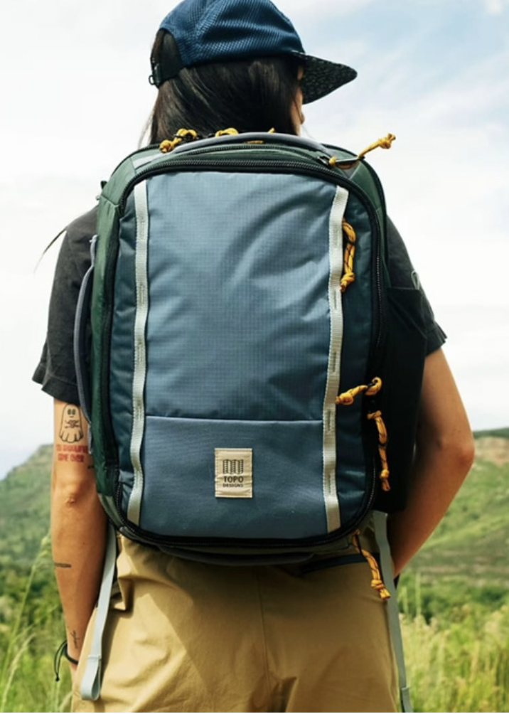 Person mit Rucksack auf einer grünen Wiese, mit Hügel im Hintergrund, trägt schwarze Kleidung, eine blaue Mütze und hat Tattoos am linken Arm.
