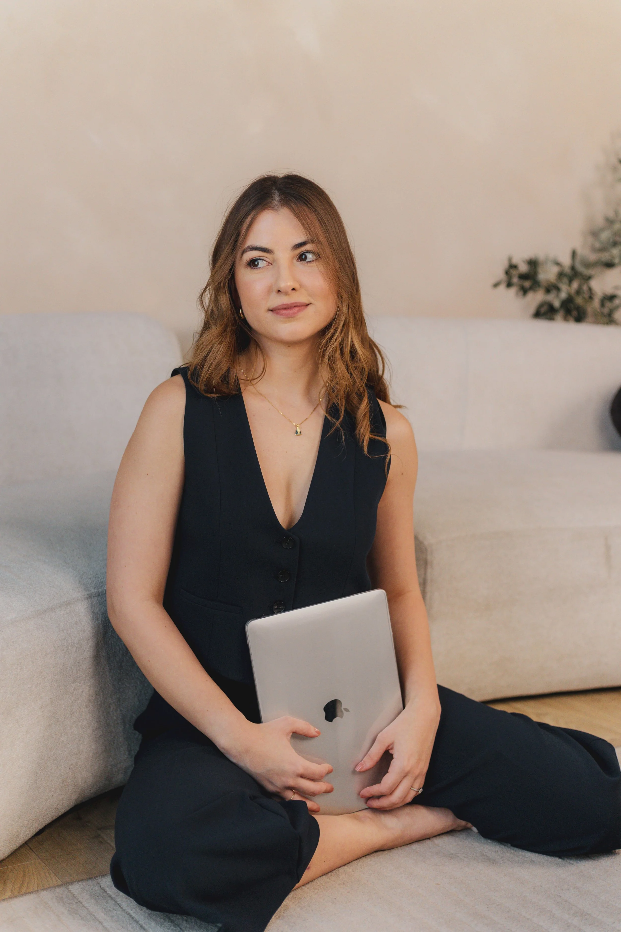 smiling at laptop, teaching how to sell Notion templates and digital products holding a silver MacBook laptop on her lap, wearing a navy sleeveless top and black pants, looking slightly to her right.