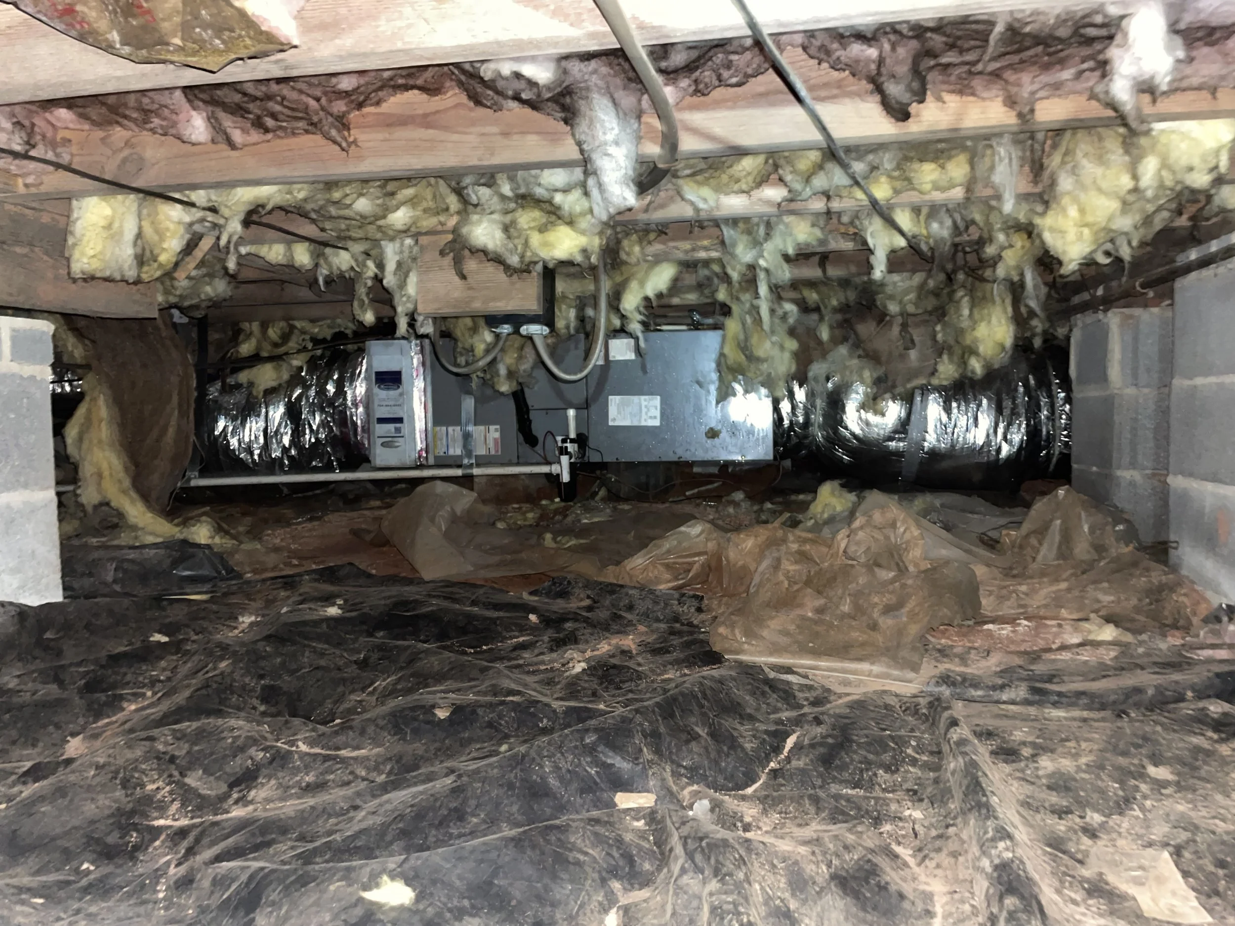Dirty crawl space with fallen insulation, damaged vapor barrier, debris and exposed HVAC equipment
