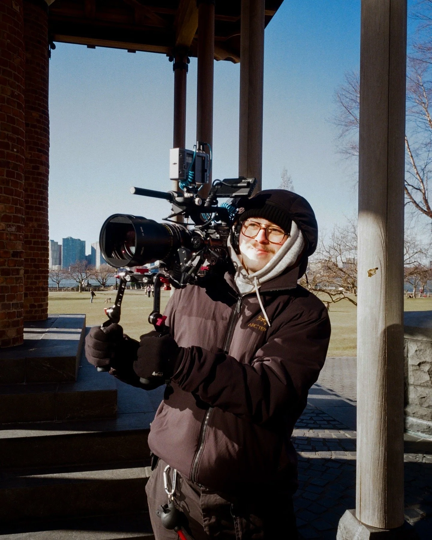 A person in outdoor winter clothing, including glasses and a beanie, holding a professional video camera on a tripod outdoors near a brick and wooden structure with a park and city skyline in the background.