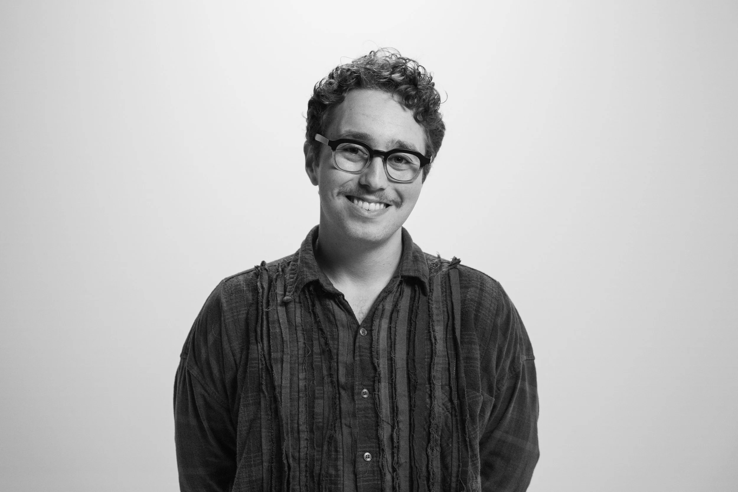 Black and white portrait of a young man with curly hair, wearing glasses and a patterned shirt, smiling against a plain white background.