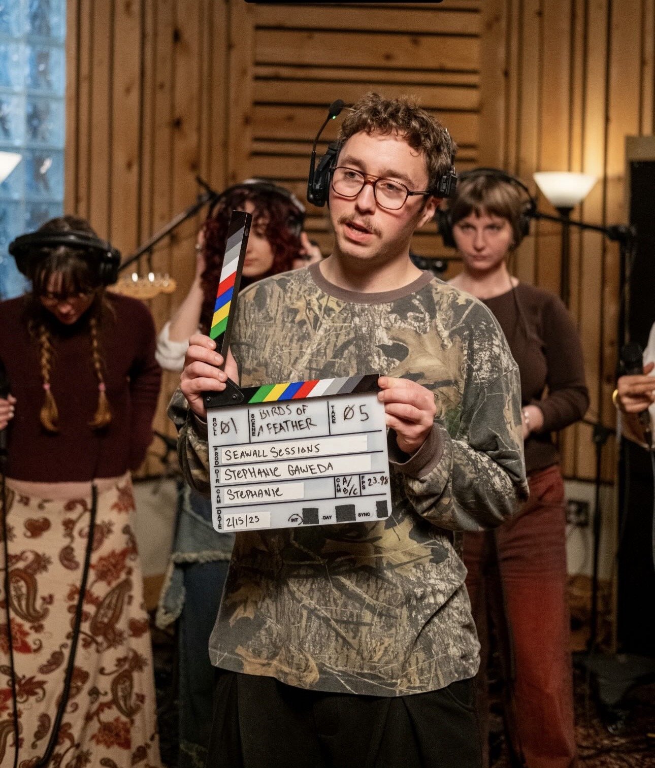 A man with glasses and headphones holding a film slate in a recording studio, with three women in the background, all surrounded by wooden paneling.
