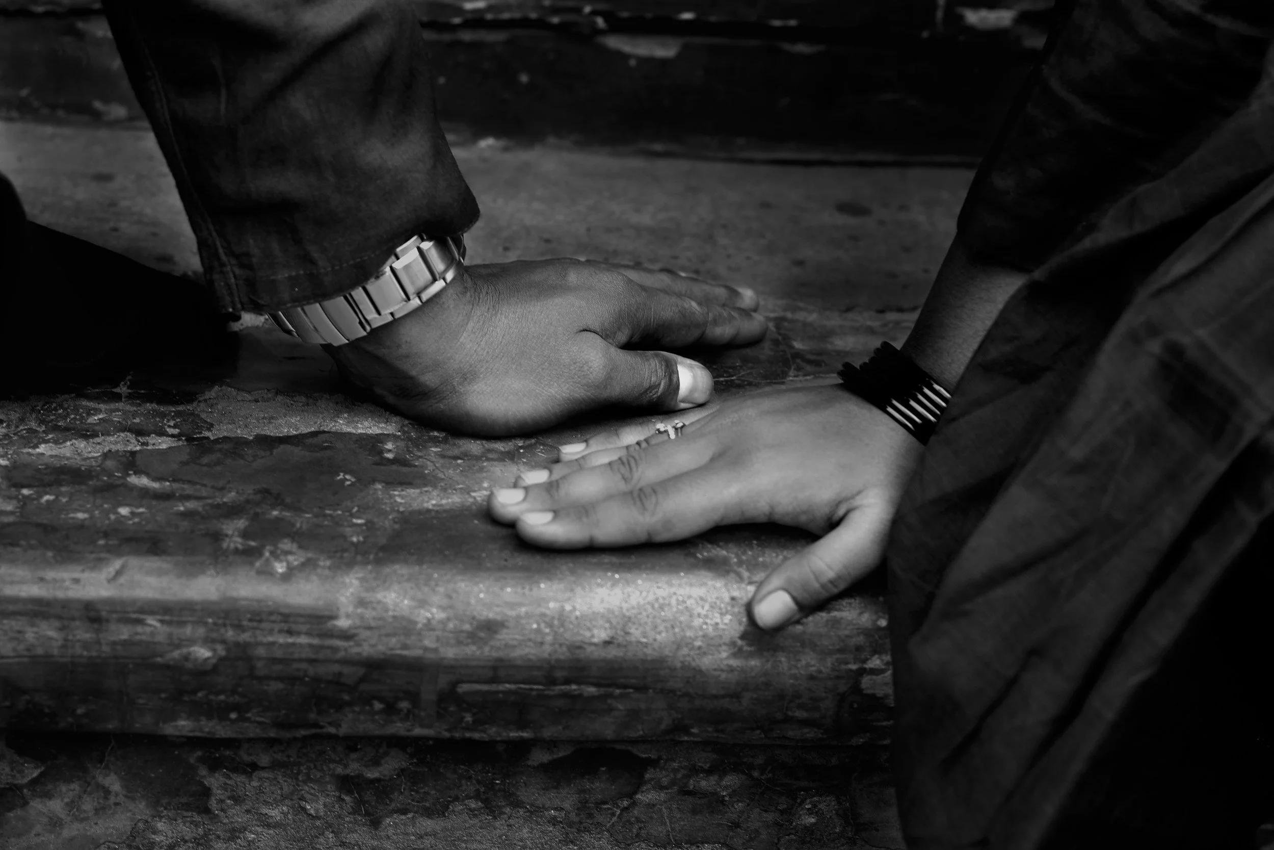 Two hands touching on a wooden surface, with one hand positioned over the other. The person wearing a watch and a ring appears to be reaching out to the other person.
