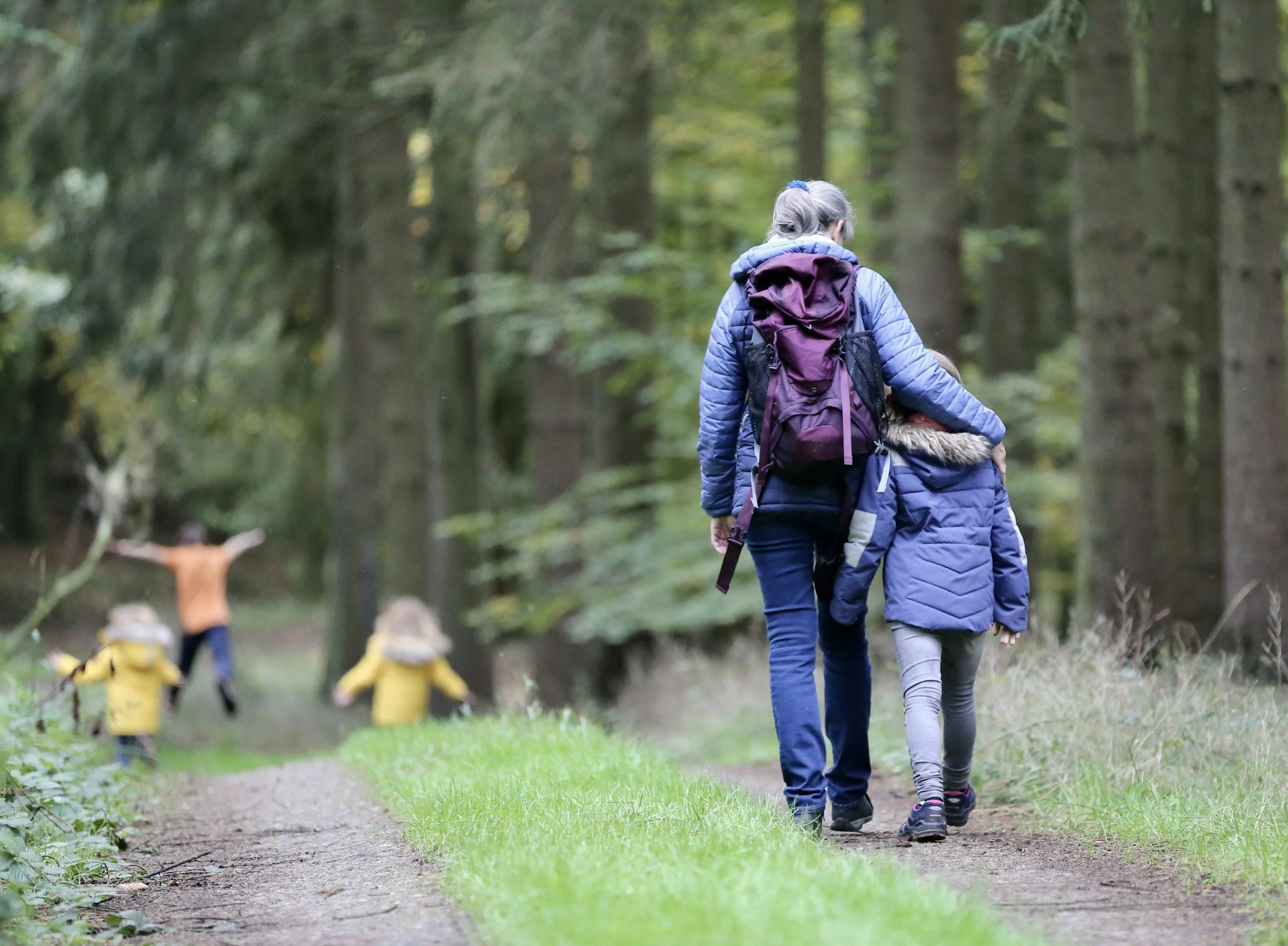 Two people, a taller woman and a smaller girl, walking in a forest on a dirt trail. They are wearing blue jackets and carrying backpacks. In the background, there are trees and a dog with a yellow raincoat and yellow boots.