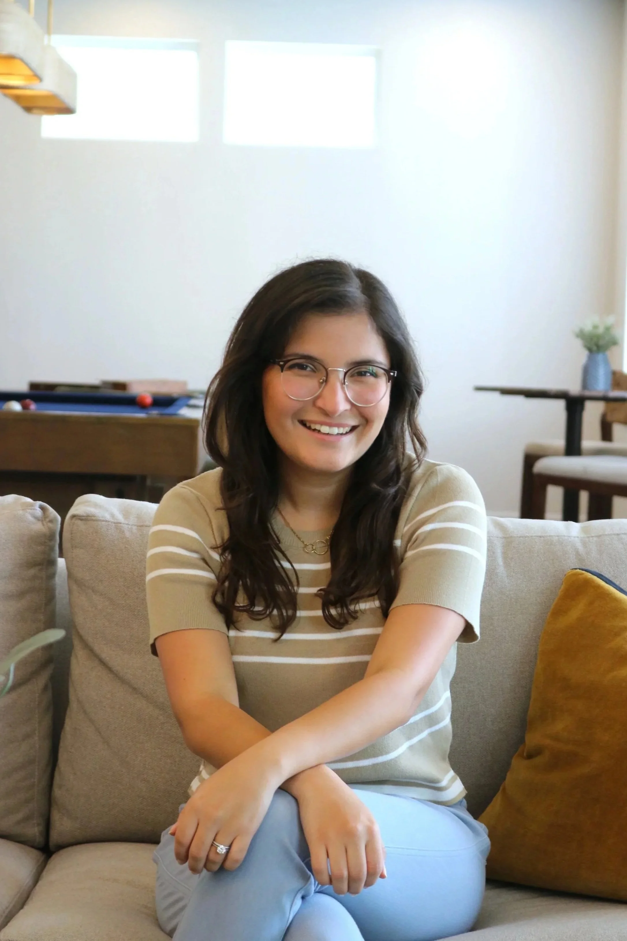 A young woman with long dark wavy hair and glasses smiling, sitting on a beige sofa crossed her arms, wearing a beige and white striped short-sleeve shirt and light blue pants, in a cozy living room with sunlight coming through two bright rectangular windows, a billiard table in the background, and a small round table with a plant on it to her right.