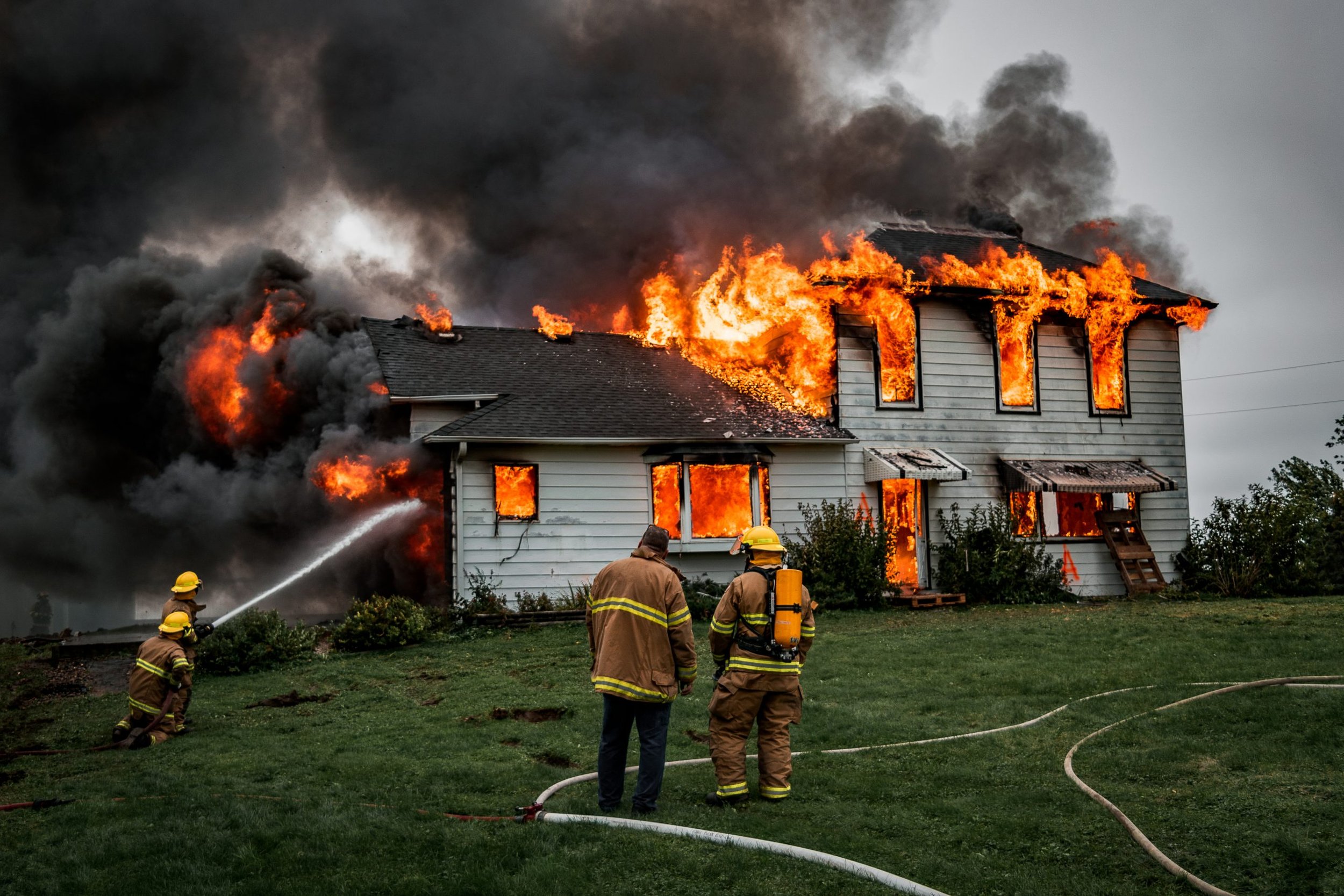 Firefighters battling a house fire with flames and thick black smoke coming from the house. The house has a two-story structure, and firefighters are spraying water onto the flames while standing on the lawn.