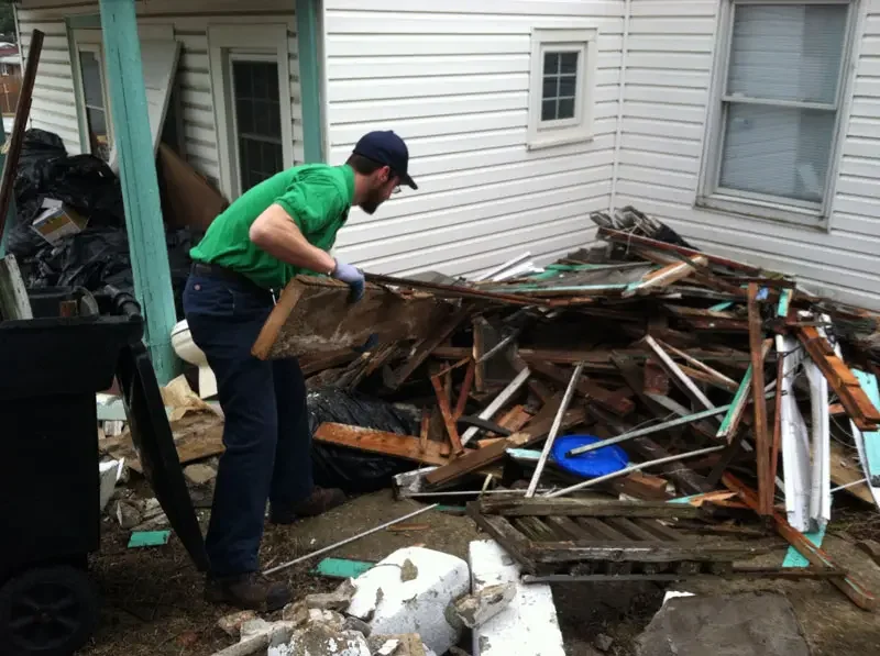 Man with gloves cleaning up debris after a house damage, using a shovel, next to a pile of wood and broken materials.