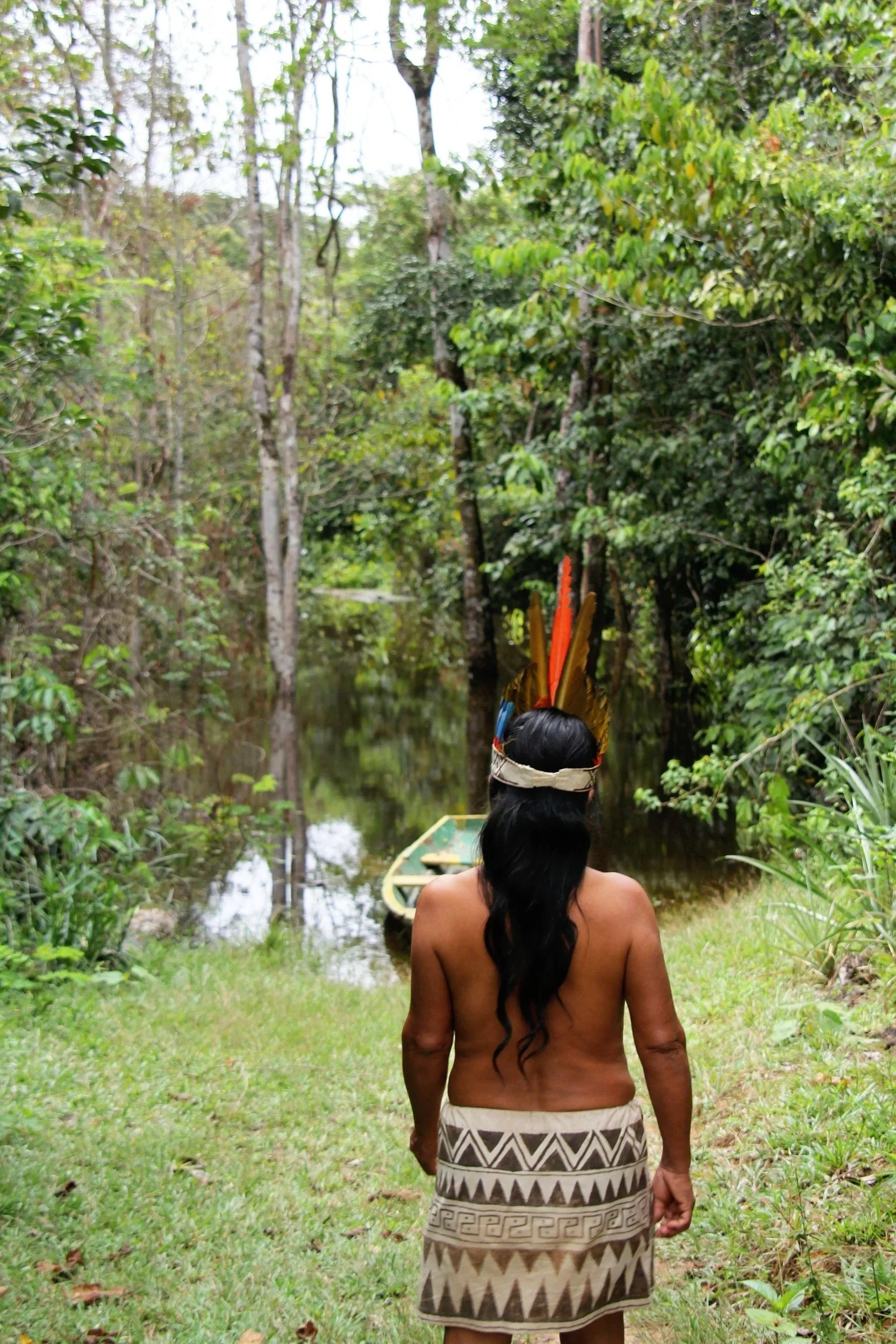 Ein Shipibo Ureinwohner steht mit dem Rücken zur Kamera vor einem Fluss im Dschungel, trägt einen Rock mit tribal Muster, hat lange schwarze Haare und einen Federkranz auf dem Kopf. Vor ihm schwimmt ein grünes Ruderboot im Wasser.