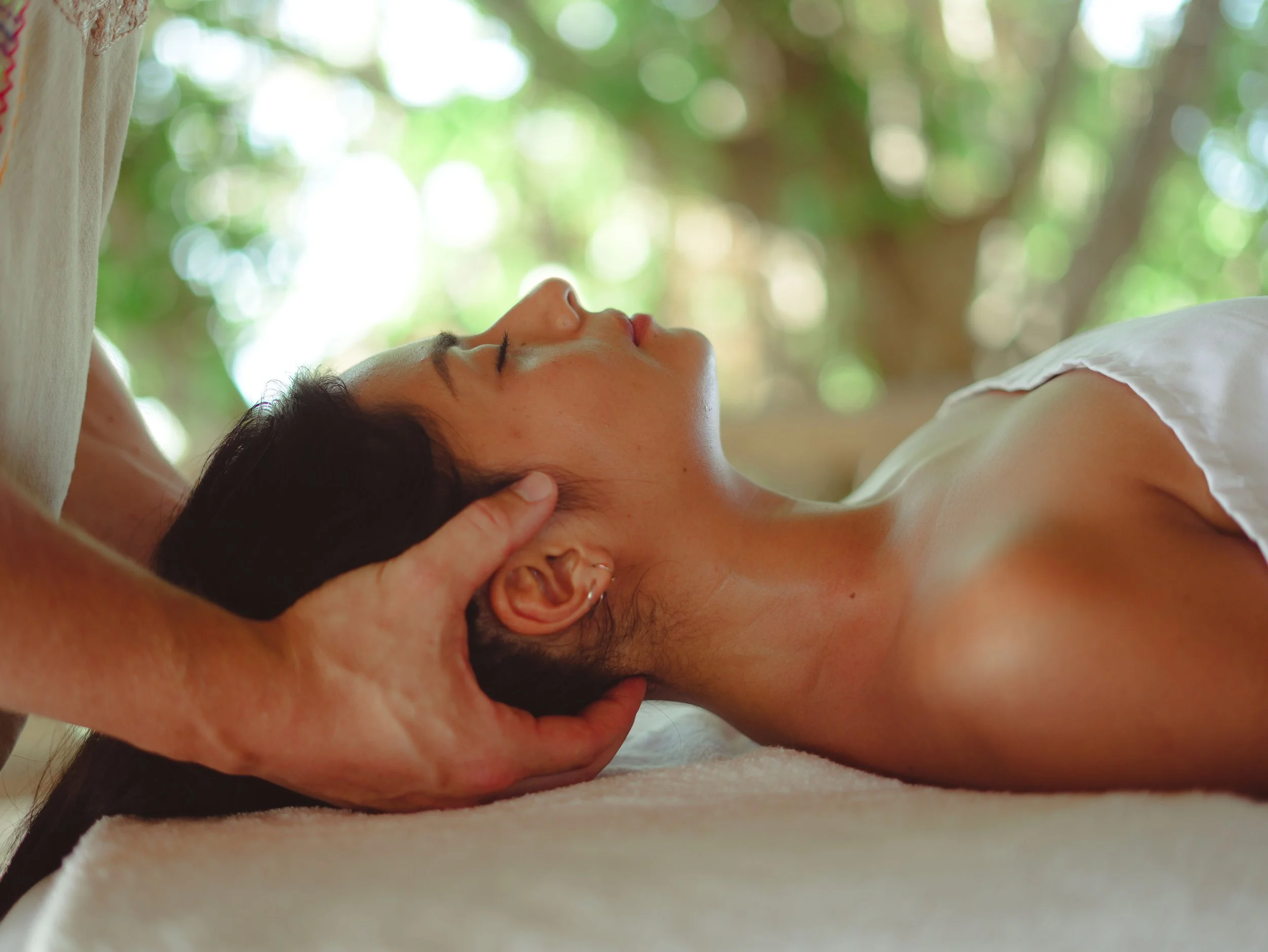 Eine junge Frau erhält auf einer Massageliege liegend eine Kopfmassage. Ihre Augen sind geschlossen, während ihr Kopf wie in einer Schale in den Händen des Masseurs liegen.