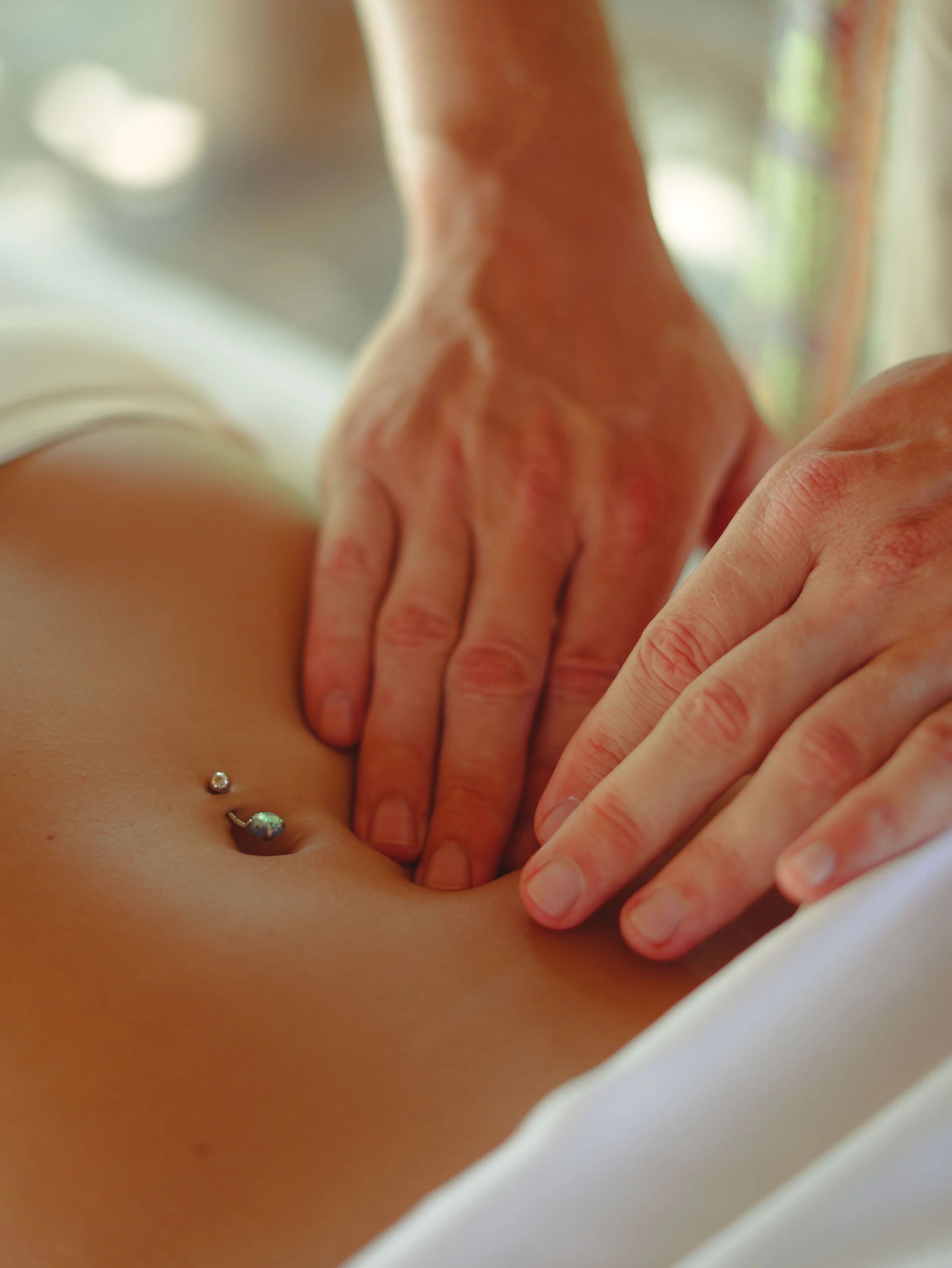 Zwei starke Hände führen eine Bauchmassage an einem Bauch mit Bauchnabelpiercing durch. Im Shiatsu auch Bauchraumerkundung genannt, soll diese Technik in der Magengegend aufgestaute Emotionen lösen.