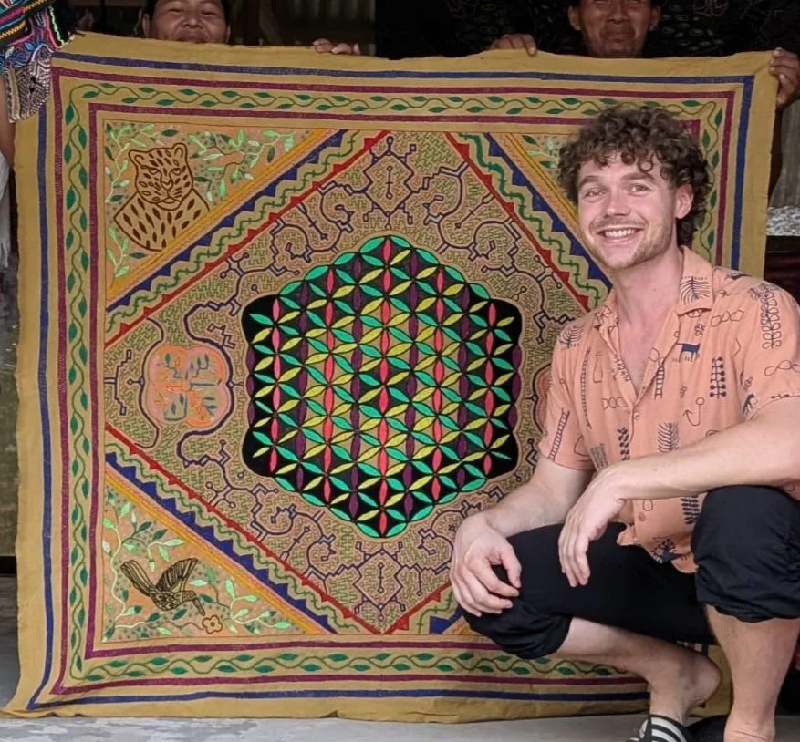 Ein lachender Mann mit lockigem Haar, der neben einem bunten, gemusterten, handgearbeiteten Stofftuch im Shipibo Stil hockt. Auf dem Stofftuch sind die Blume des Lebens, symmetrische Ayahuasca Muster, sowie ein Jaguar und ein Kolibri zu sehen.