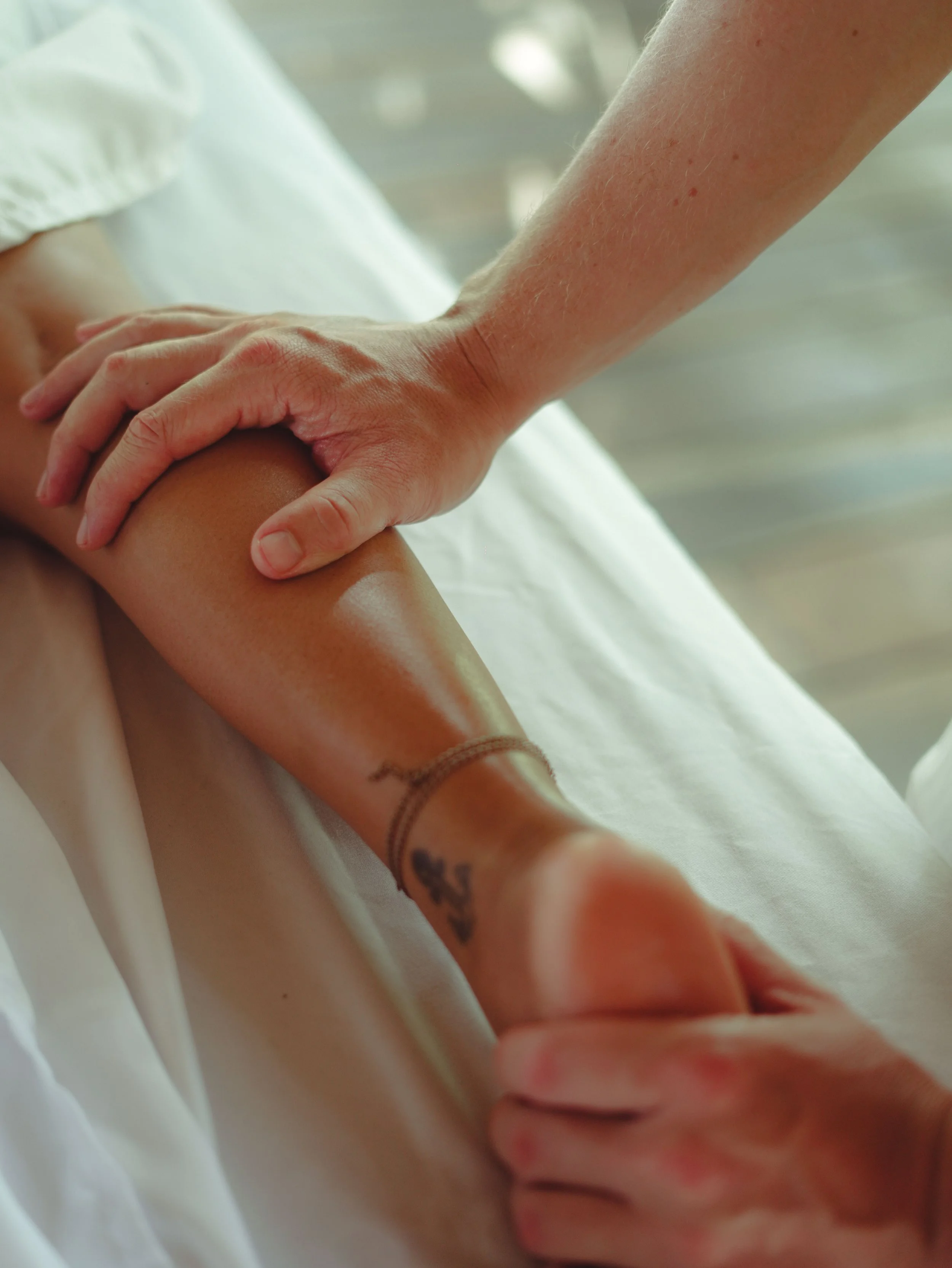 Wadenmassage bei einem femininen Bein mit Anker Tattoo und Fußkettchen. Die eine Hand des Masseurs hält den Fuß, während die andere die Wade massiert und Wadenreflexzonen stimuliert.